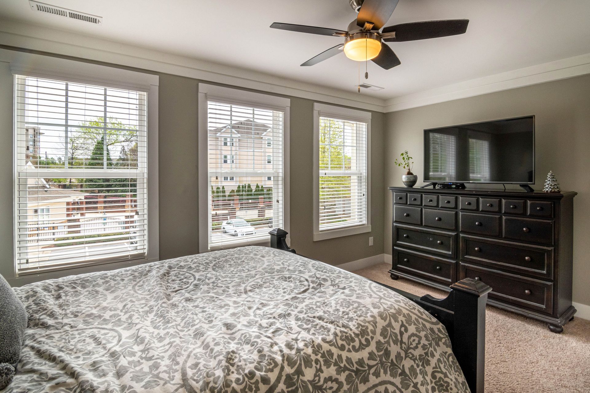 Bedroom with gray walls, patterned bedspread, three windows, ceiling fan, and dark dresser with TV.