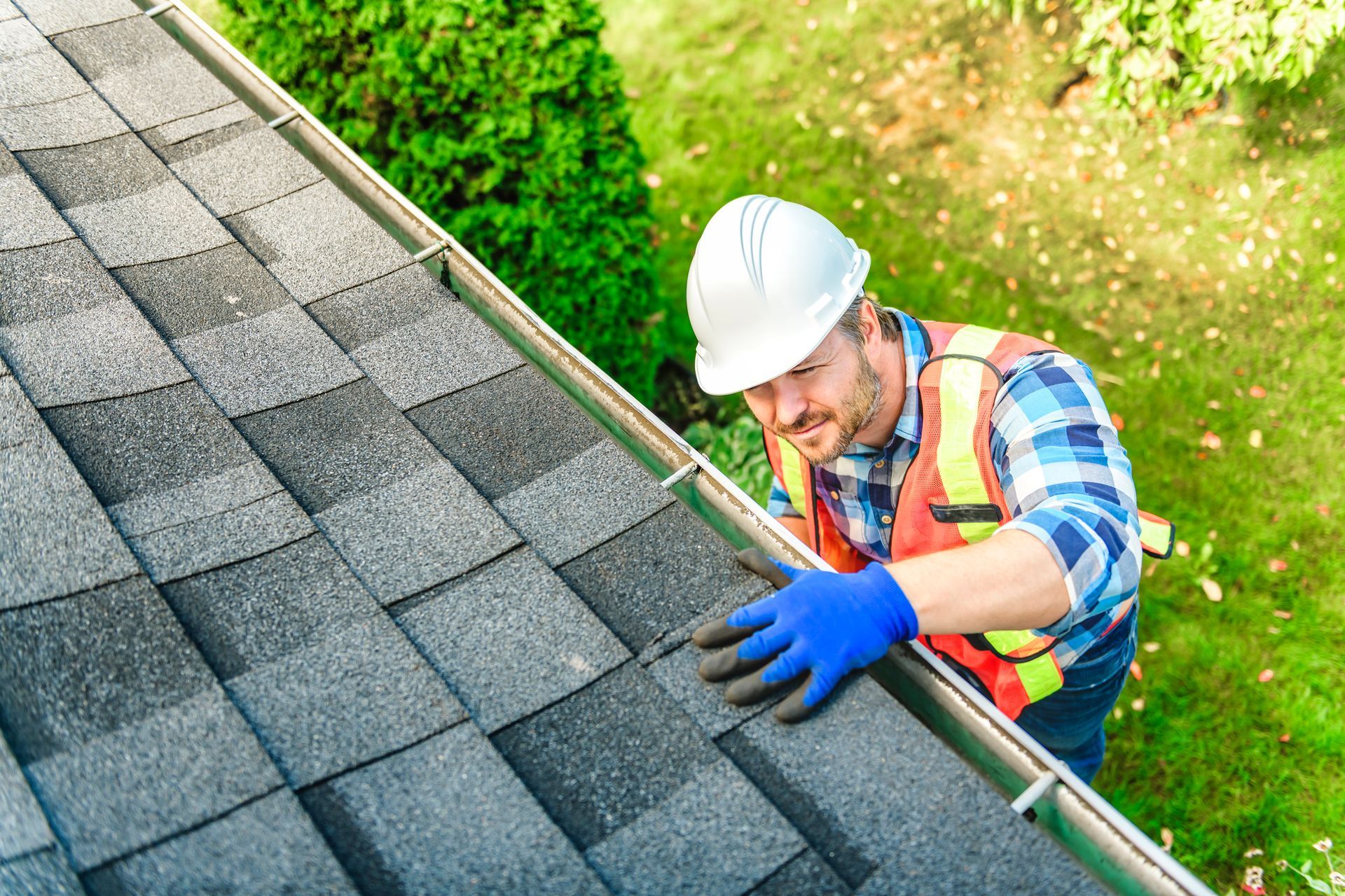 Roofer in safety gear inspecting shingles and gutter on a rooftop; greenery in background.