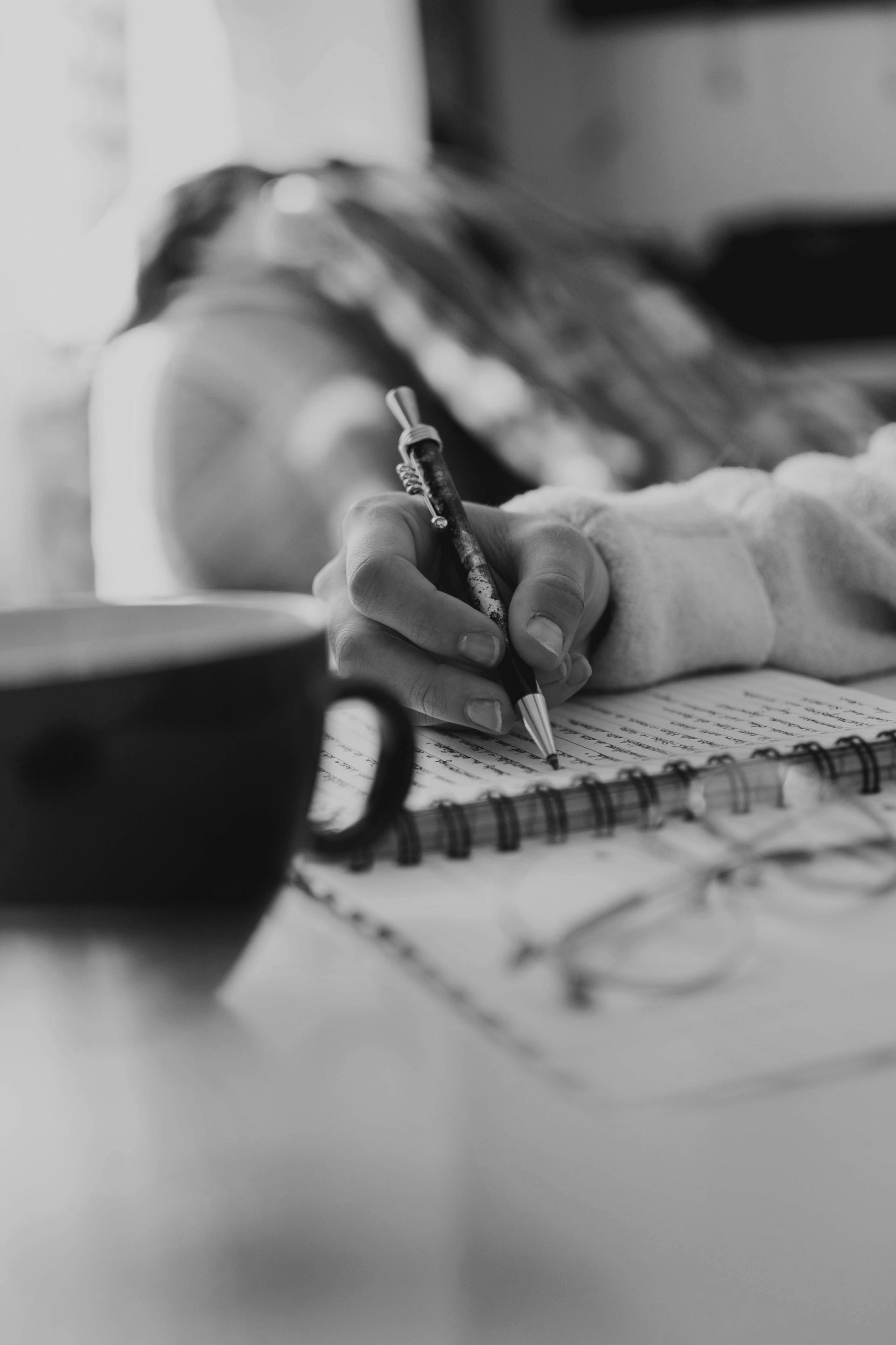 Person writing in a notebook with a pen, coffee mug in the foreground.