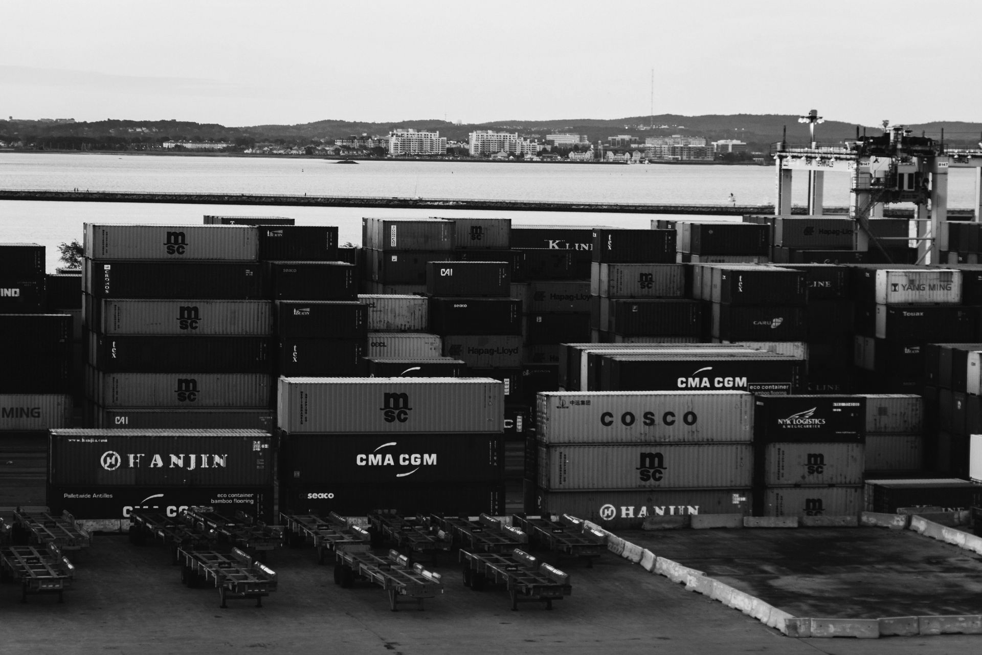 Shipping containers stacked at a port, with water and a distant cityscape in the background.