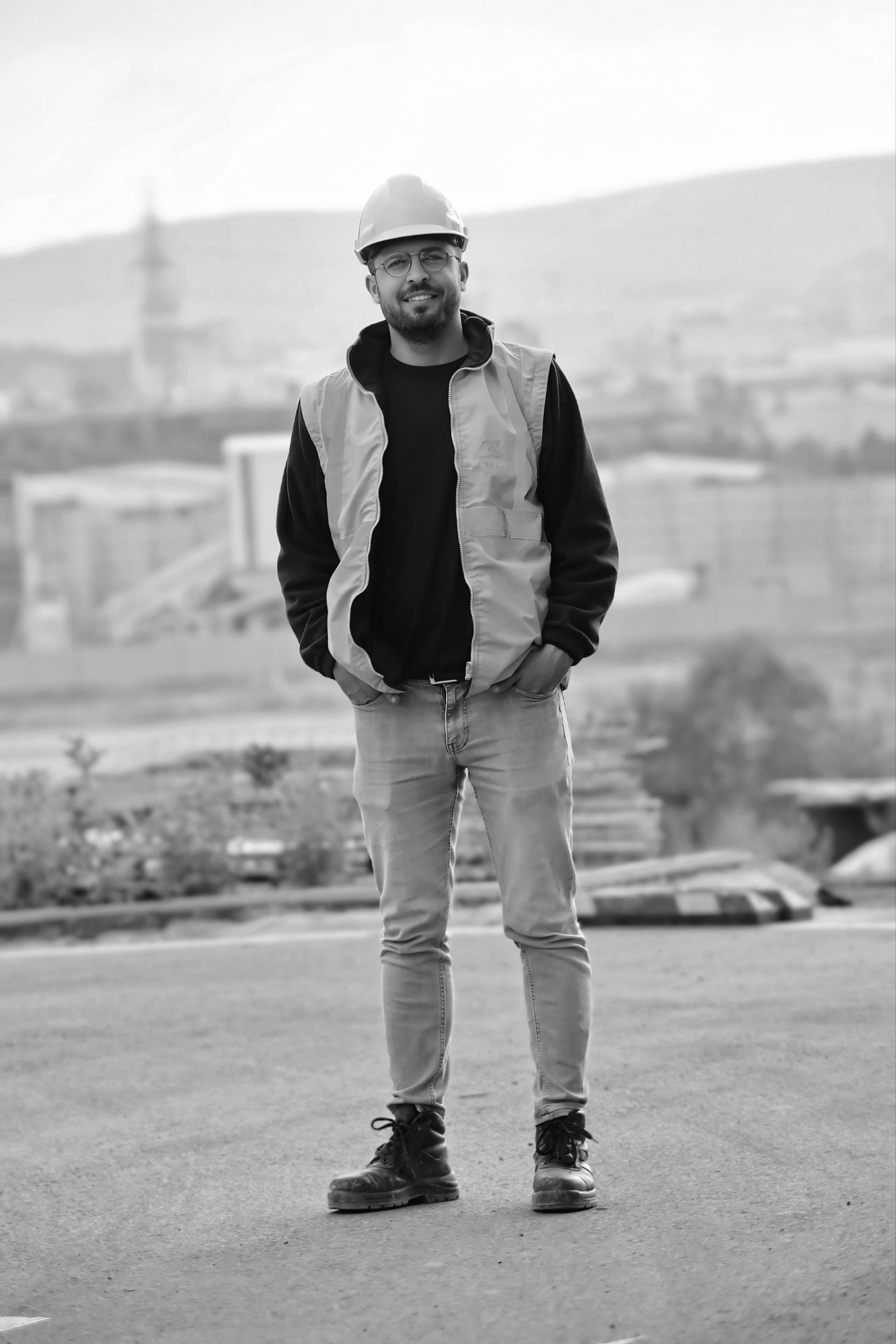 Man in hard hat and vest, standing outdoors, hands in pockets, industrial background.