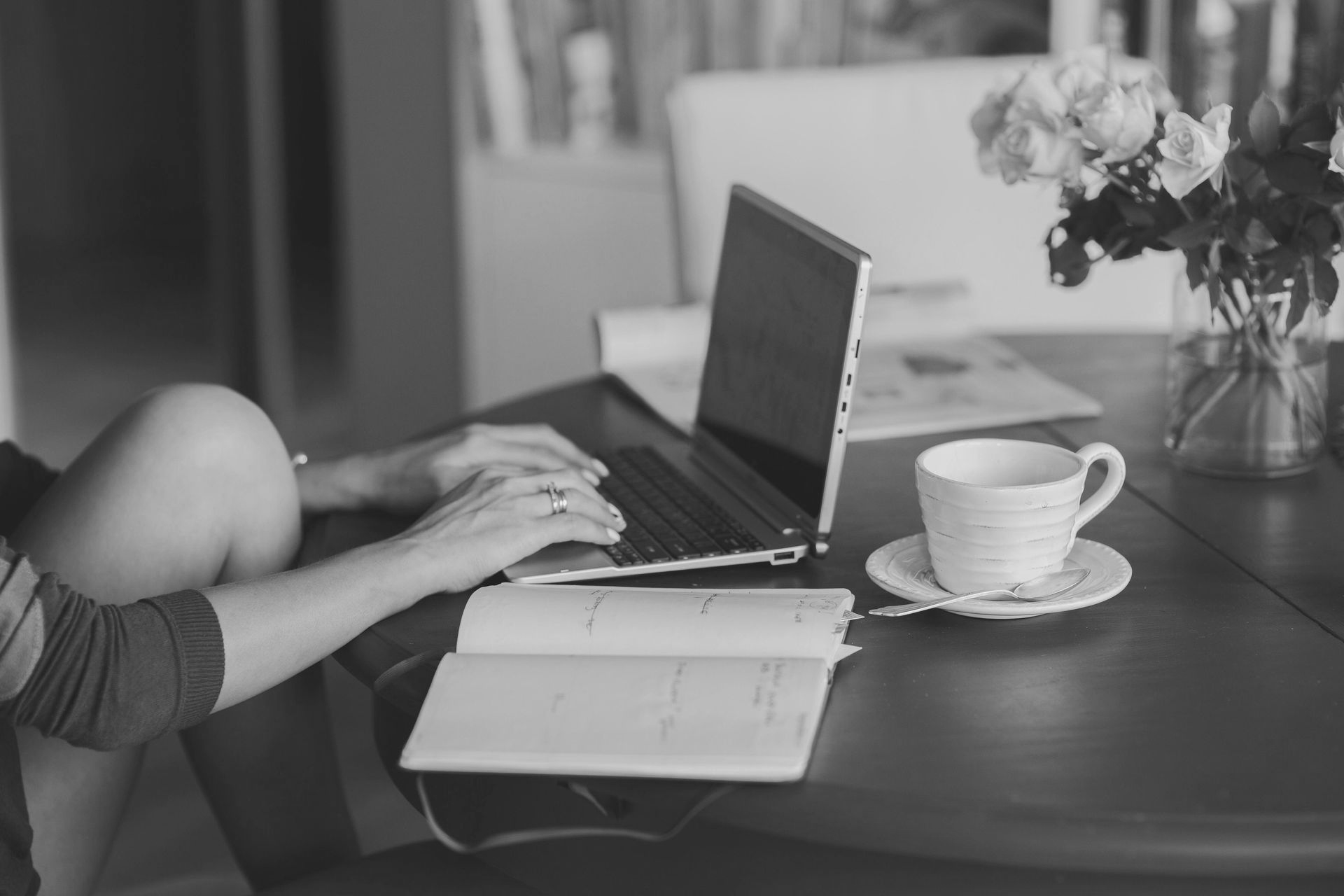 Woman typing on a laptop at a table, with coffee cup, open notebook, and flowers.