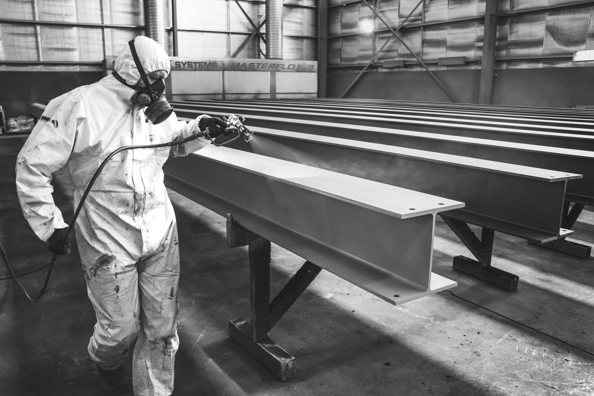 Person in protective suit spraying metal beams. Industrial setting.