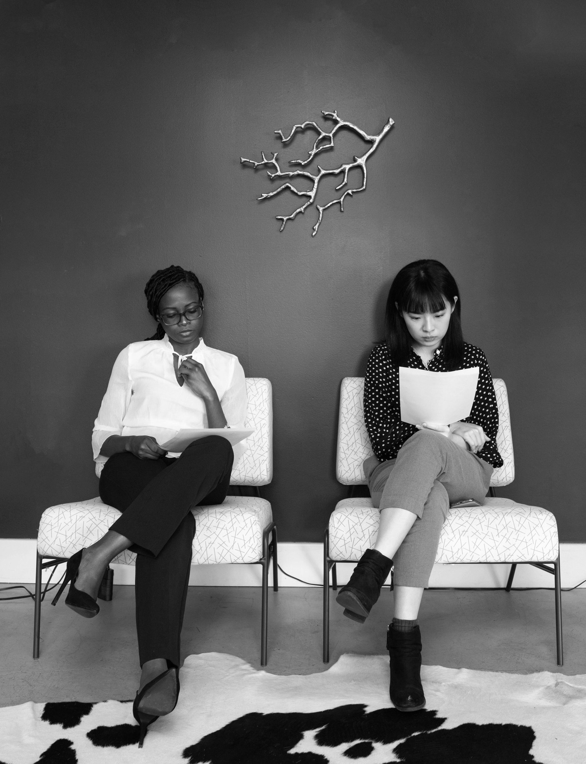 Two people seated in chairs, holding papers, looking down, in a waiting area.