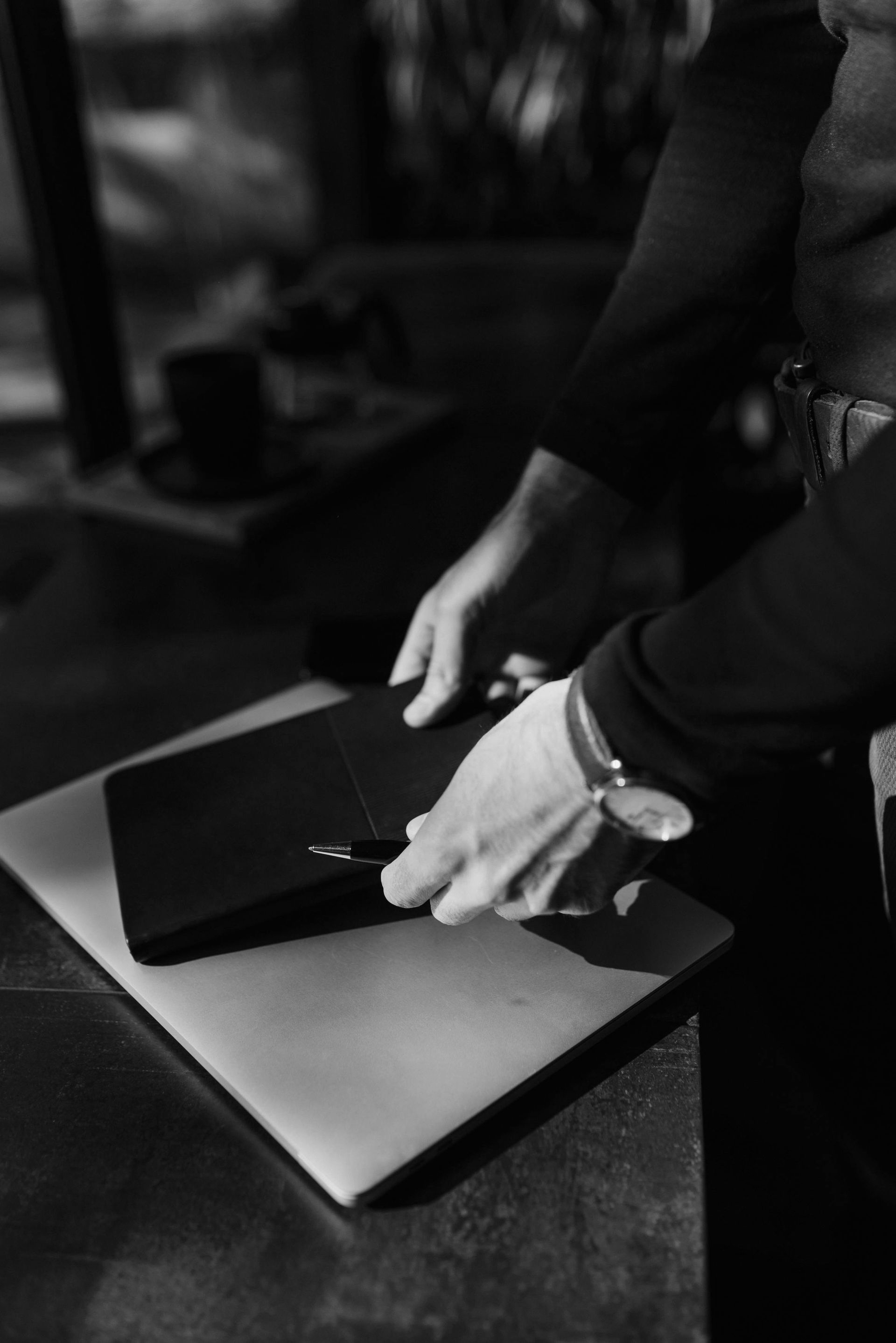Person placing a black notebook on top of a laptop, both on a dark surface.