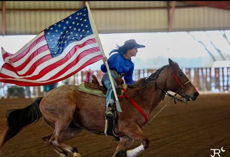 Cowboy riding a galloping horse while holding a large American flag in an indoor arena