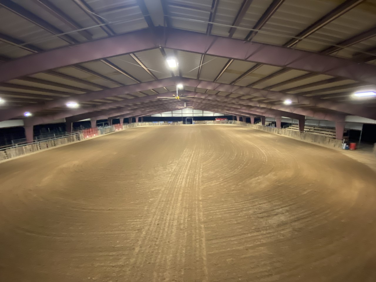 Indoor riding arena with a sandy floor and arched metal roof, viewed from the center.