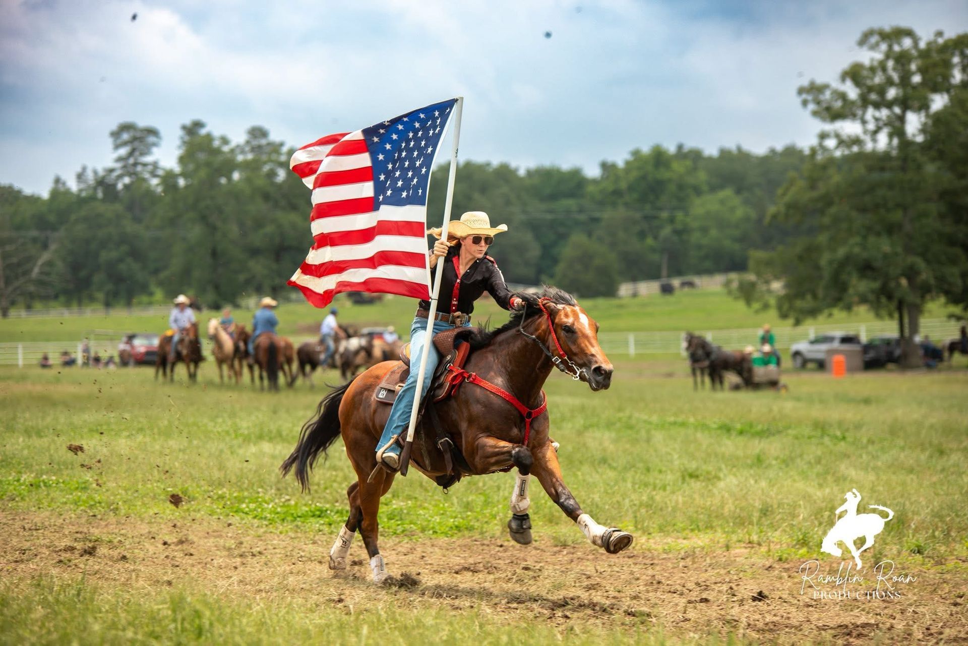 Rider on a galloping horse carries a large American flag in a muddy field, with riders in the background