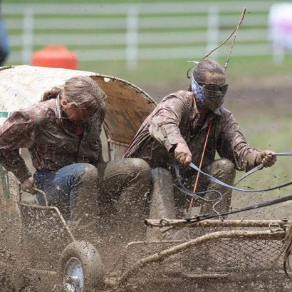 Two mud-covered riders racing a cart through a muddy track, spraying mud everywhere