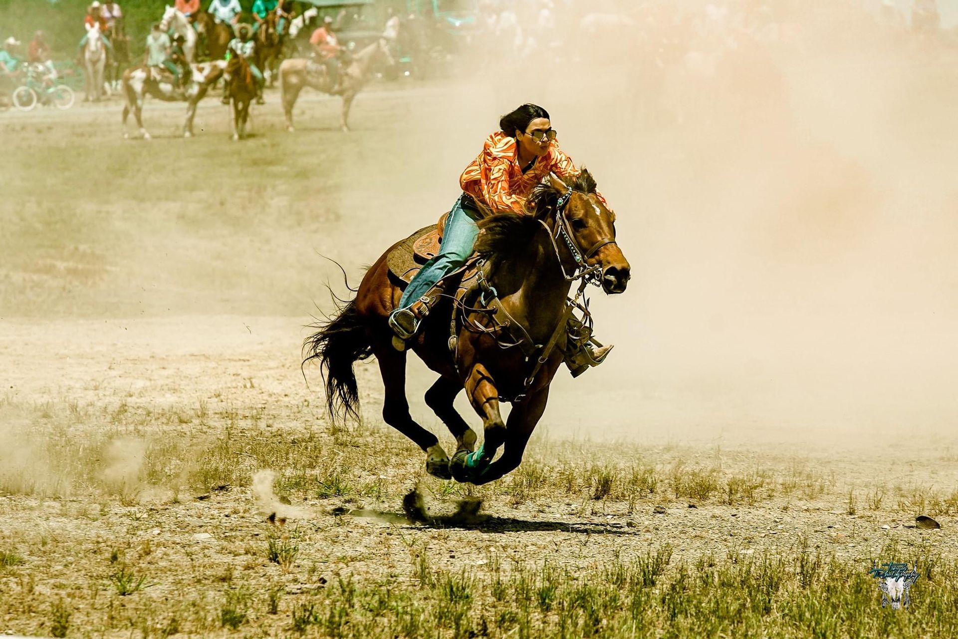 Rodeo rider in orange shirt on bucking black horse amid dusty arena, with other riders in the background