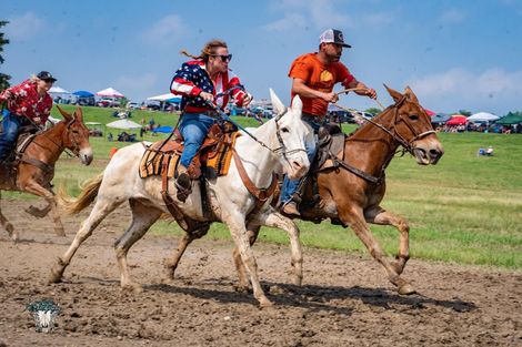Cowboys race horses across a muddy field at an outdoor rodeo.