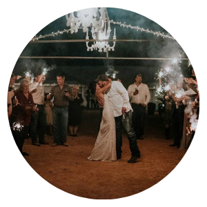 Couple kissing at a dimly lit indoor celebration with guests, chandeliers, and sparklers.