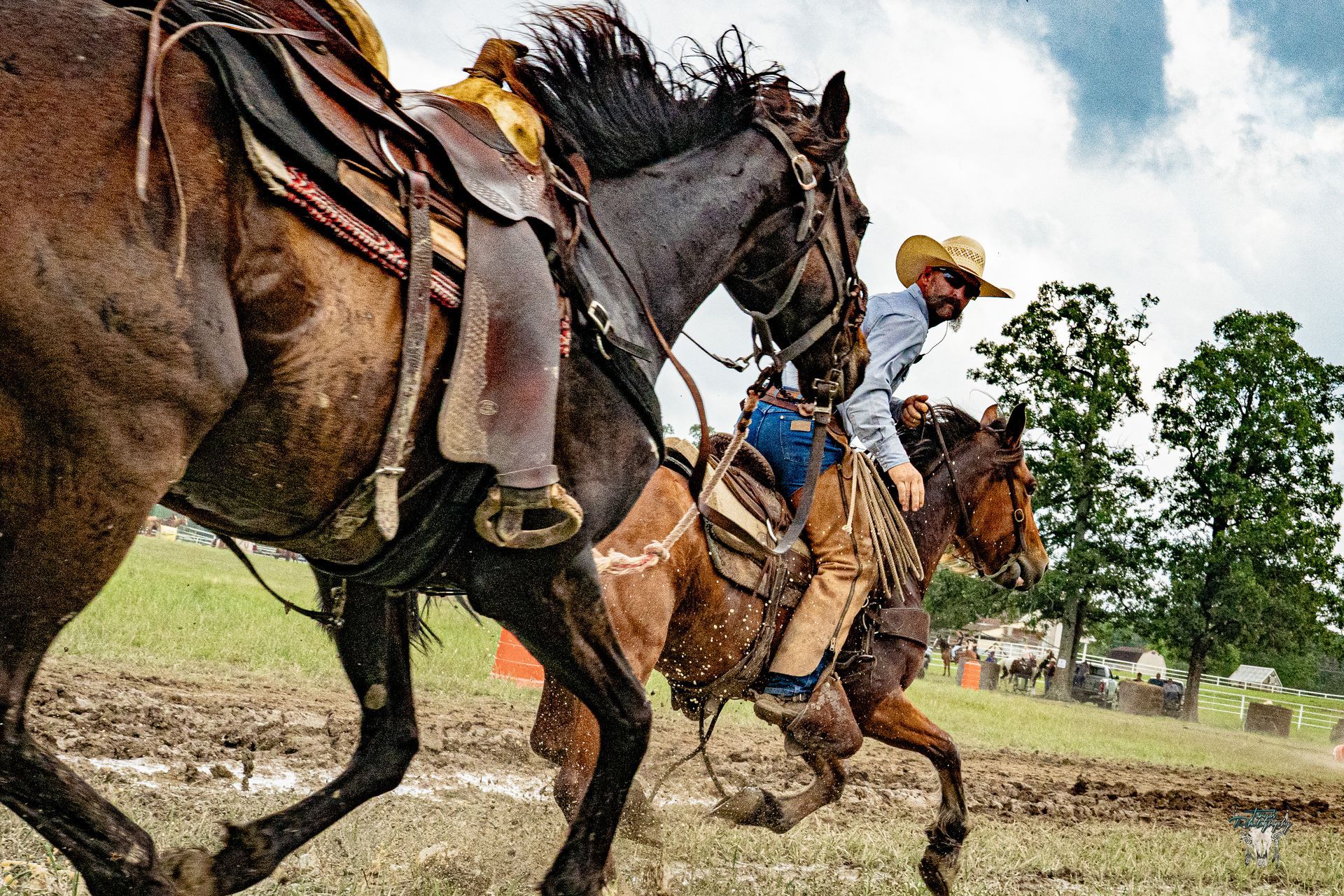 Two riders on galloping horses in a muddy field, with one in a cowboy hat