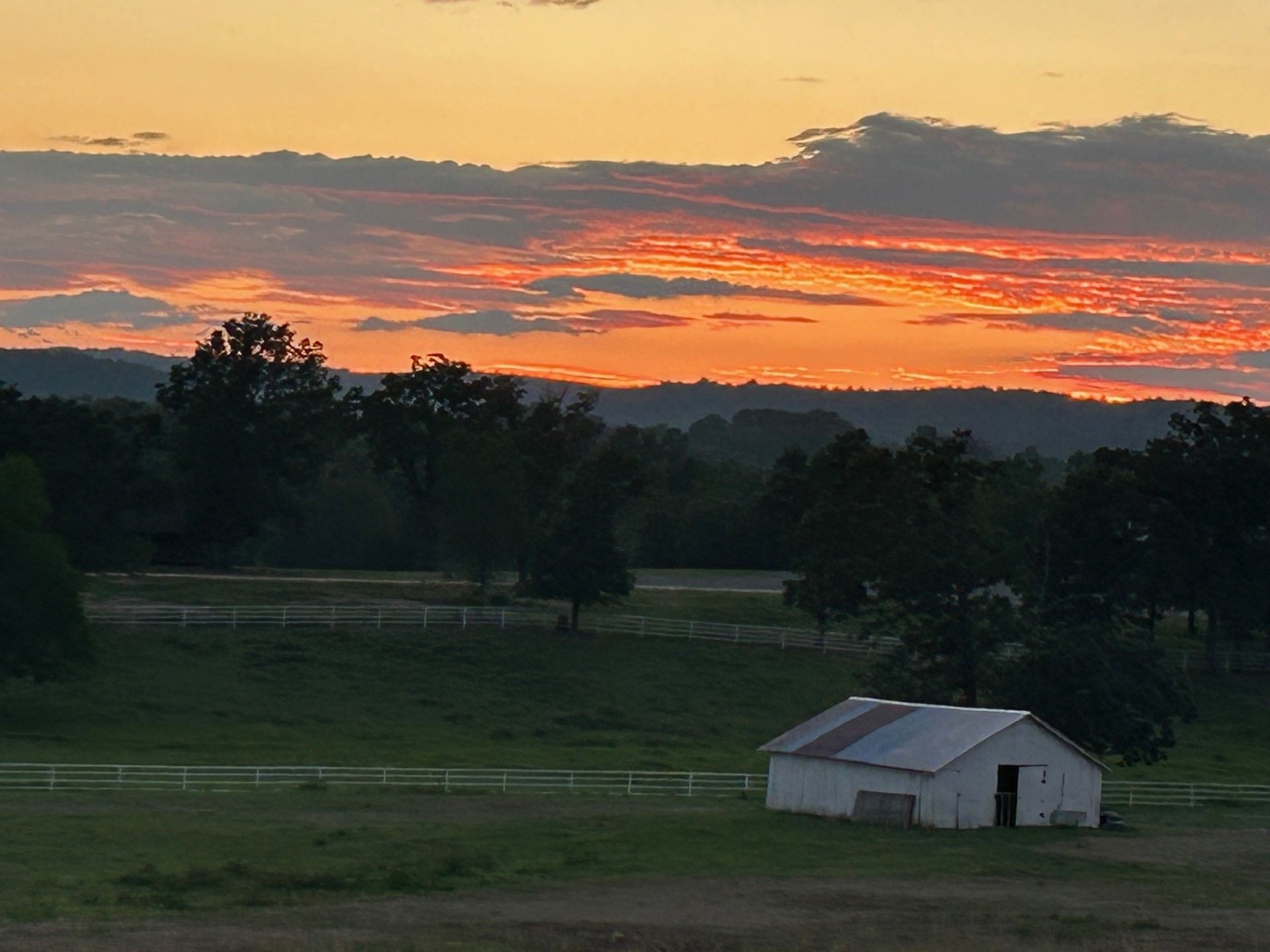 Sunset over a grassy field with a small white barn and dark trees under orange clouds