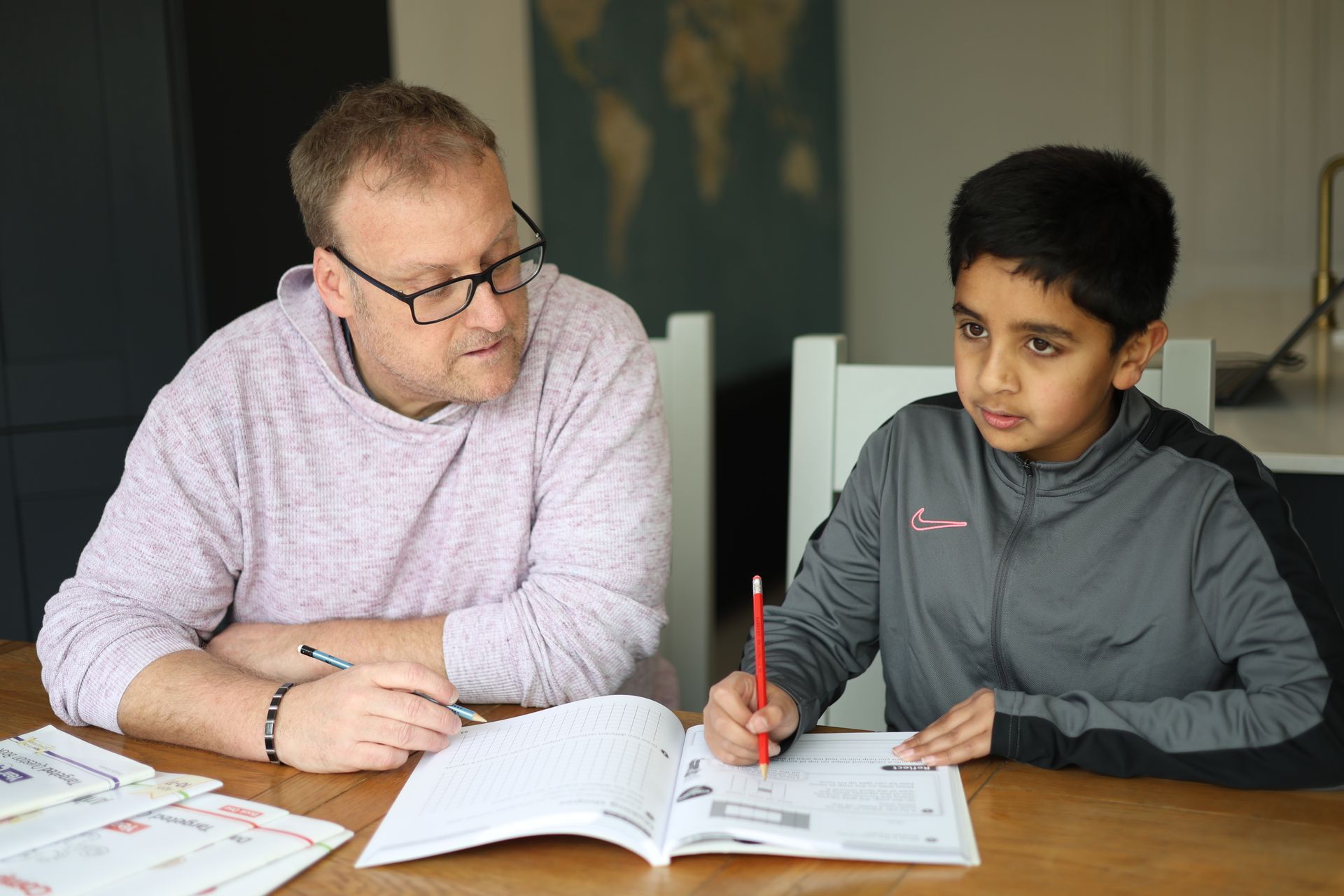 Man in glasses helping a boy with homework at a table. Both are looking at the book.