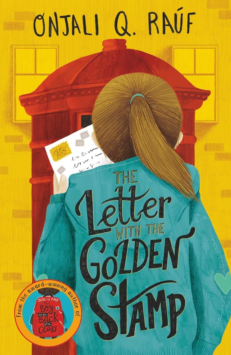 A girl in a blue jacket is holding a letter in front of a post box.