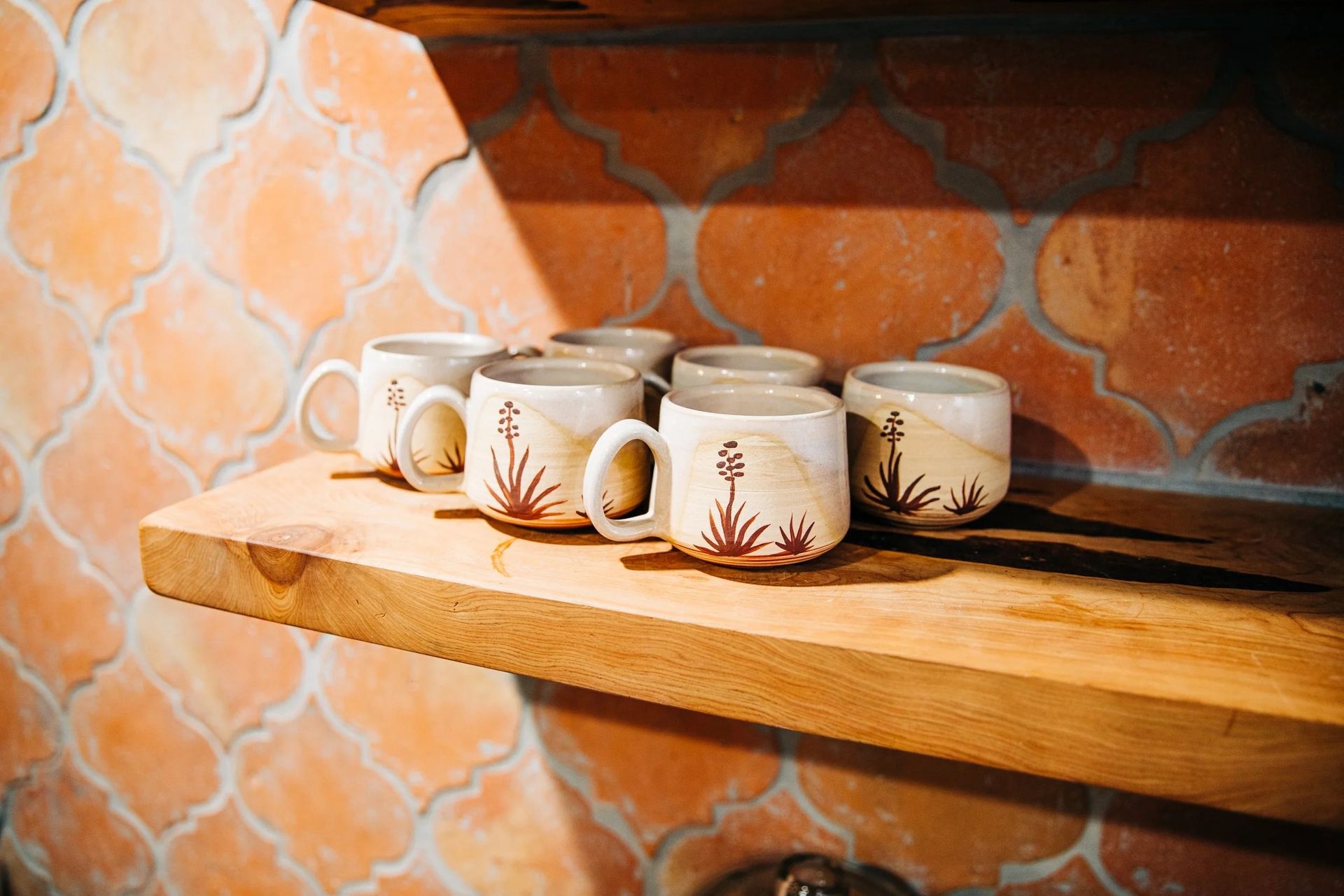 Six ceramic mugs with floral designs on a wooden shelf against an orange tiled wall.