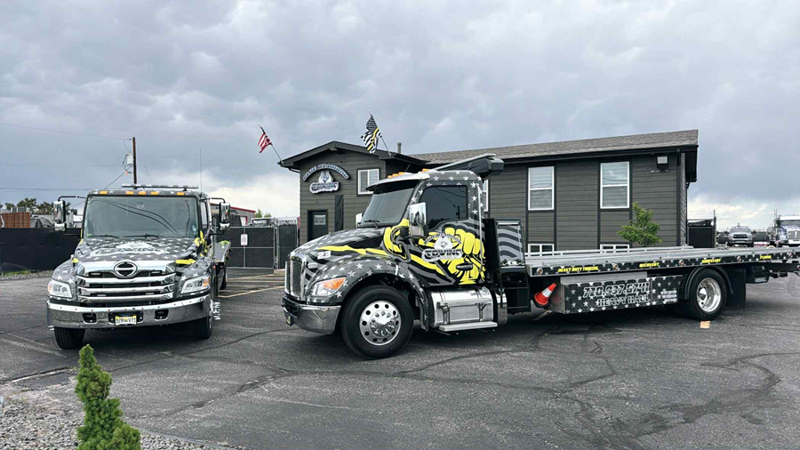Tow trucks parked in front of a building with an American flag, both trucks have unique designs.