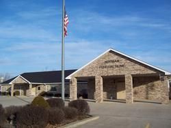A tan brick funeral home building under a clear blue sky, with a tall flagpole flying an American flag in front.
