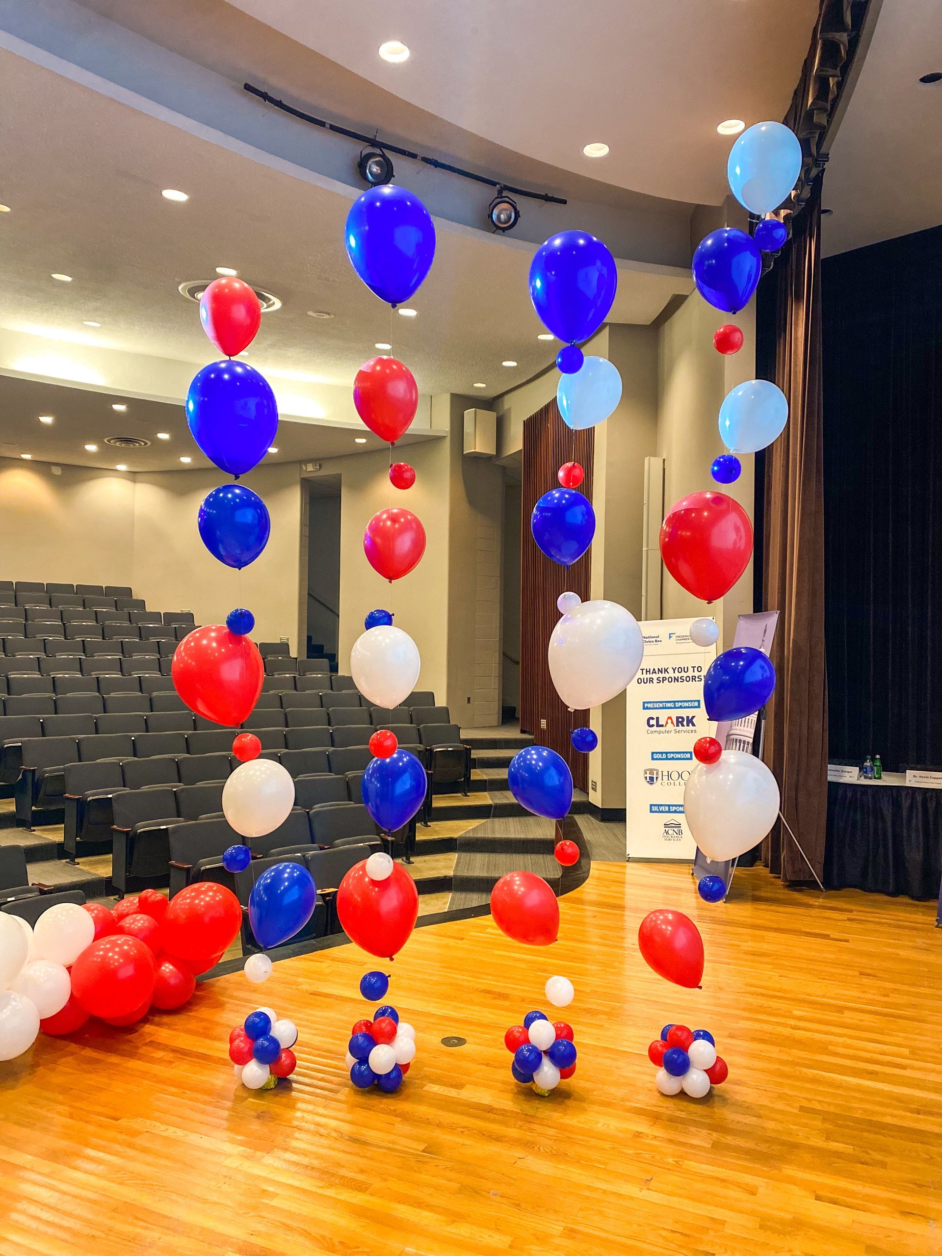 Red , white , and blue balloons are hanging from the ceiling in an auditorium.