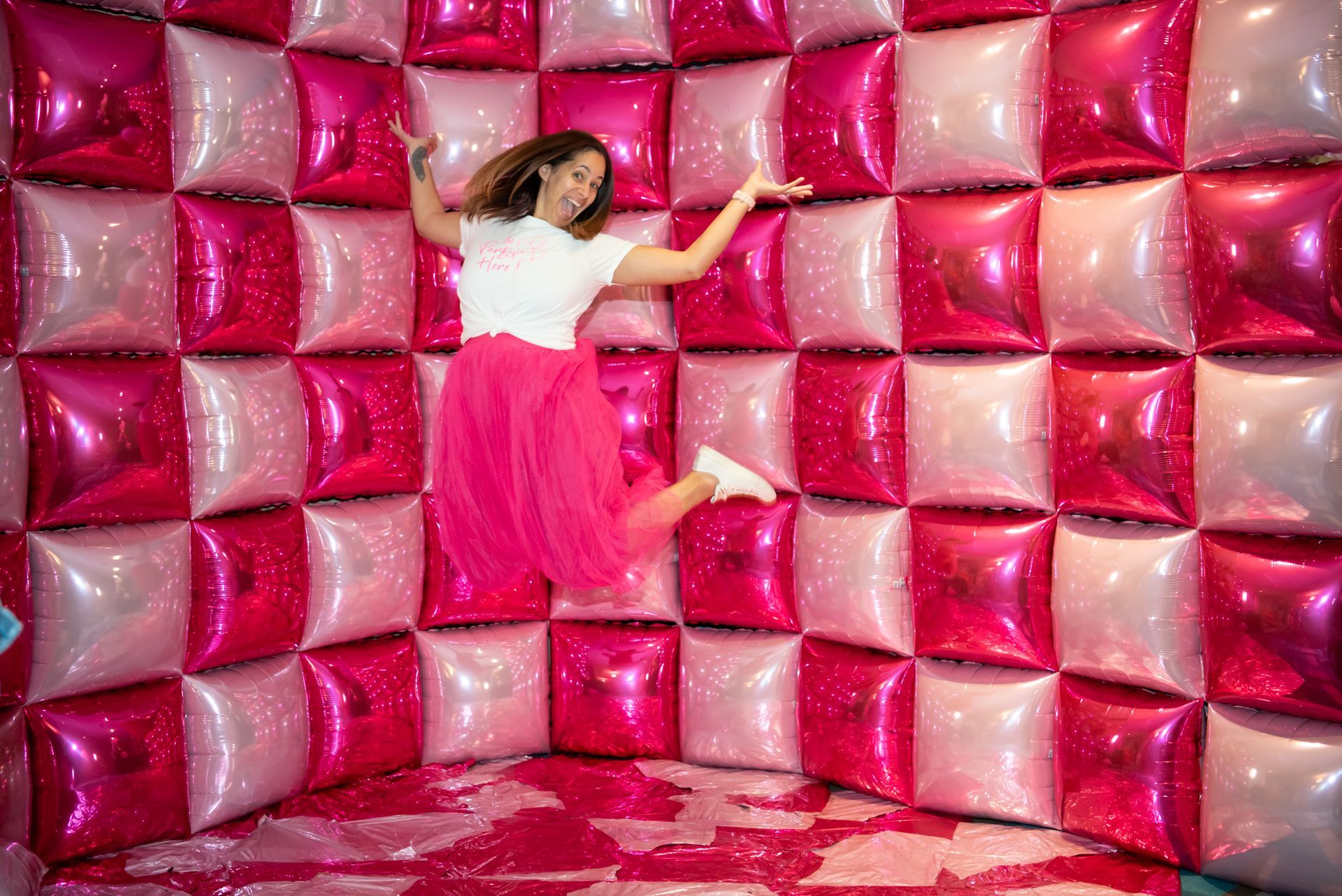 A woman is jumping in the air in a room filled with pink and white balloons.