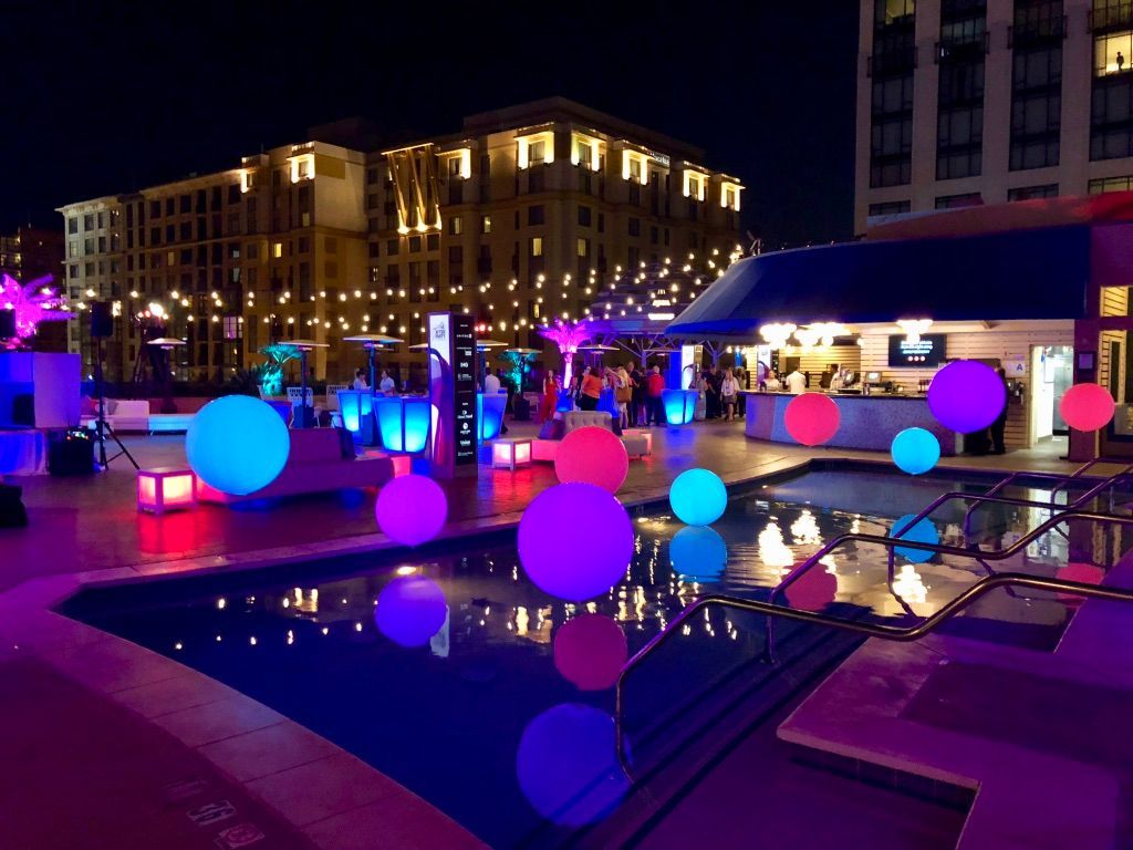 Nighttime pool party with glowing orbs, colorful lights, bar, and buildings in background.
