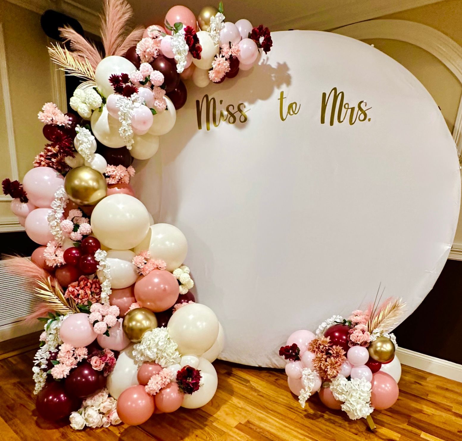 A large white circle surrounded by pink and gold balloons and flowers.
