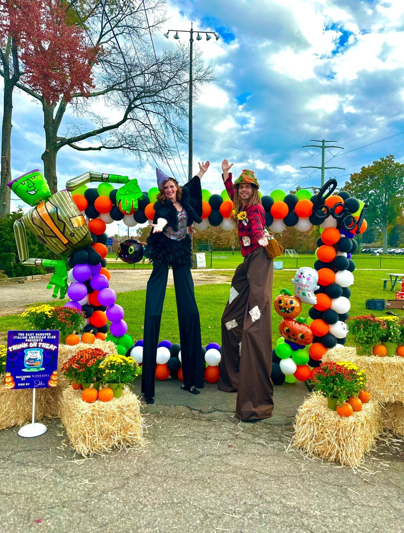 Two women on stilts are posing for a picture in front of a balloon arch.