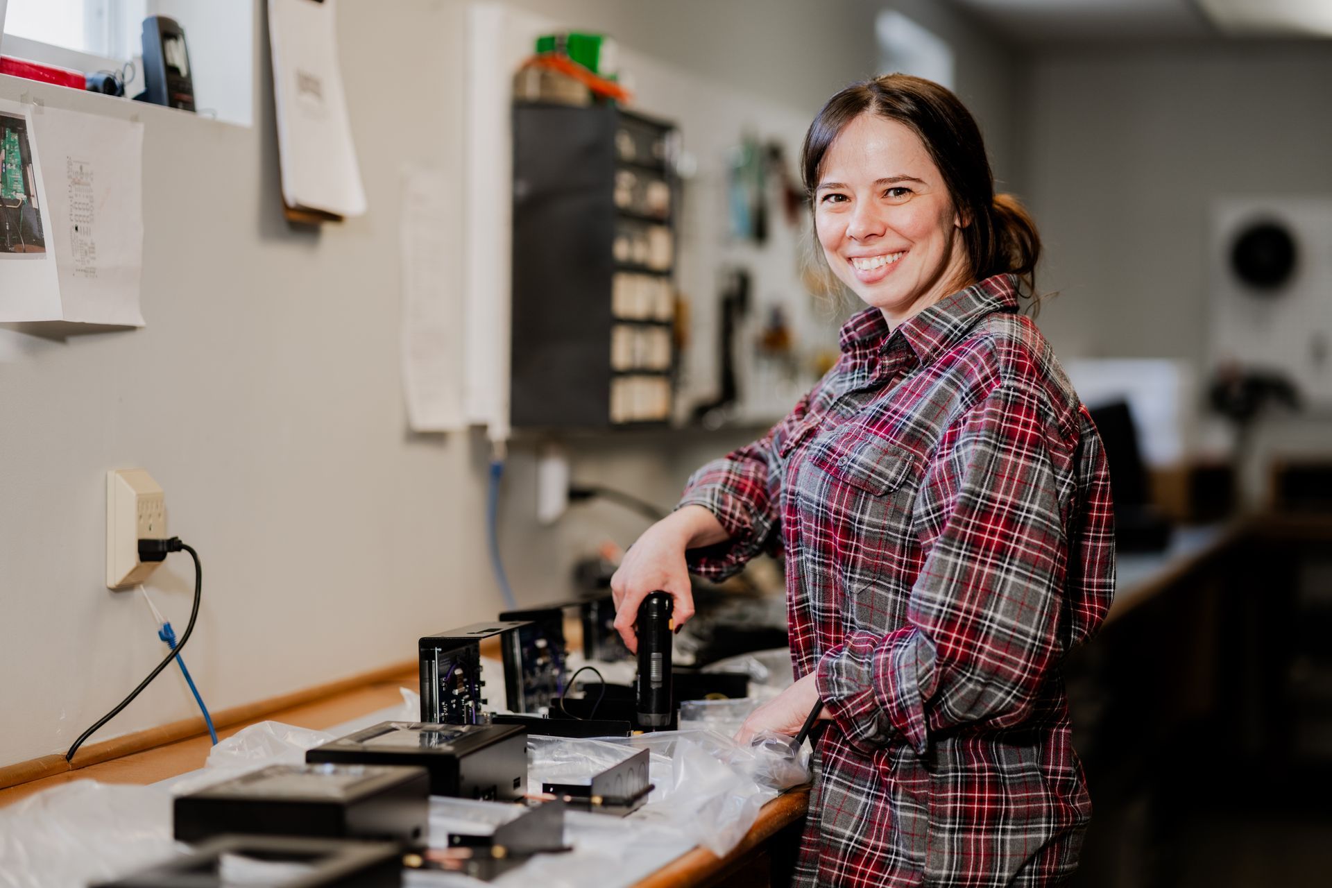 Woman in plaid shirt smiles while working on machinery at a workbench.