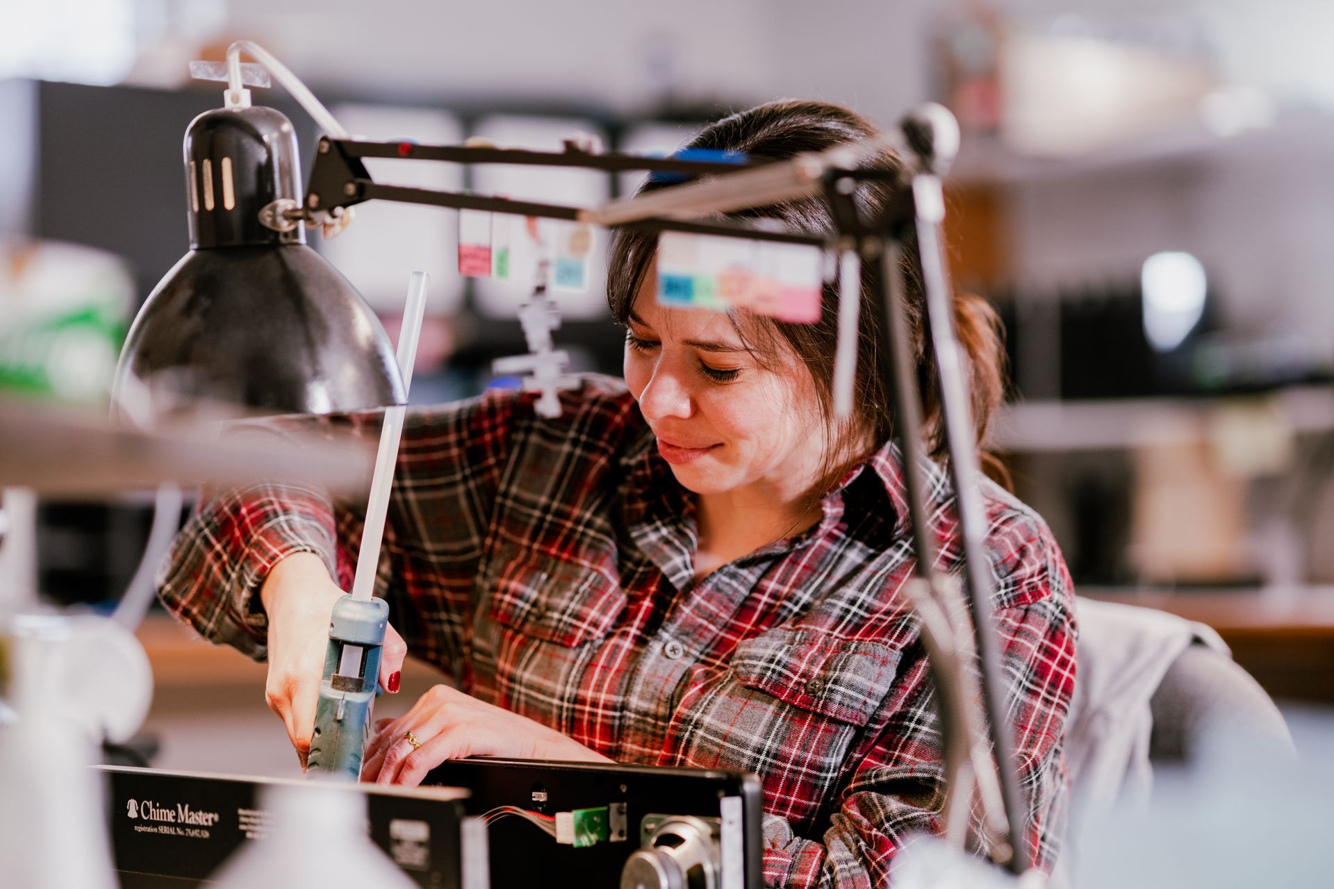 Woman in plaid shirt works at a lab bench, focused, using a tool with a light overhead.