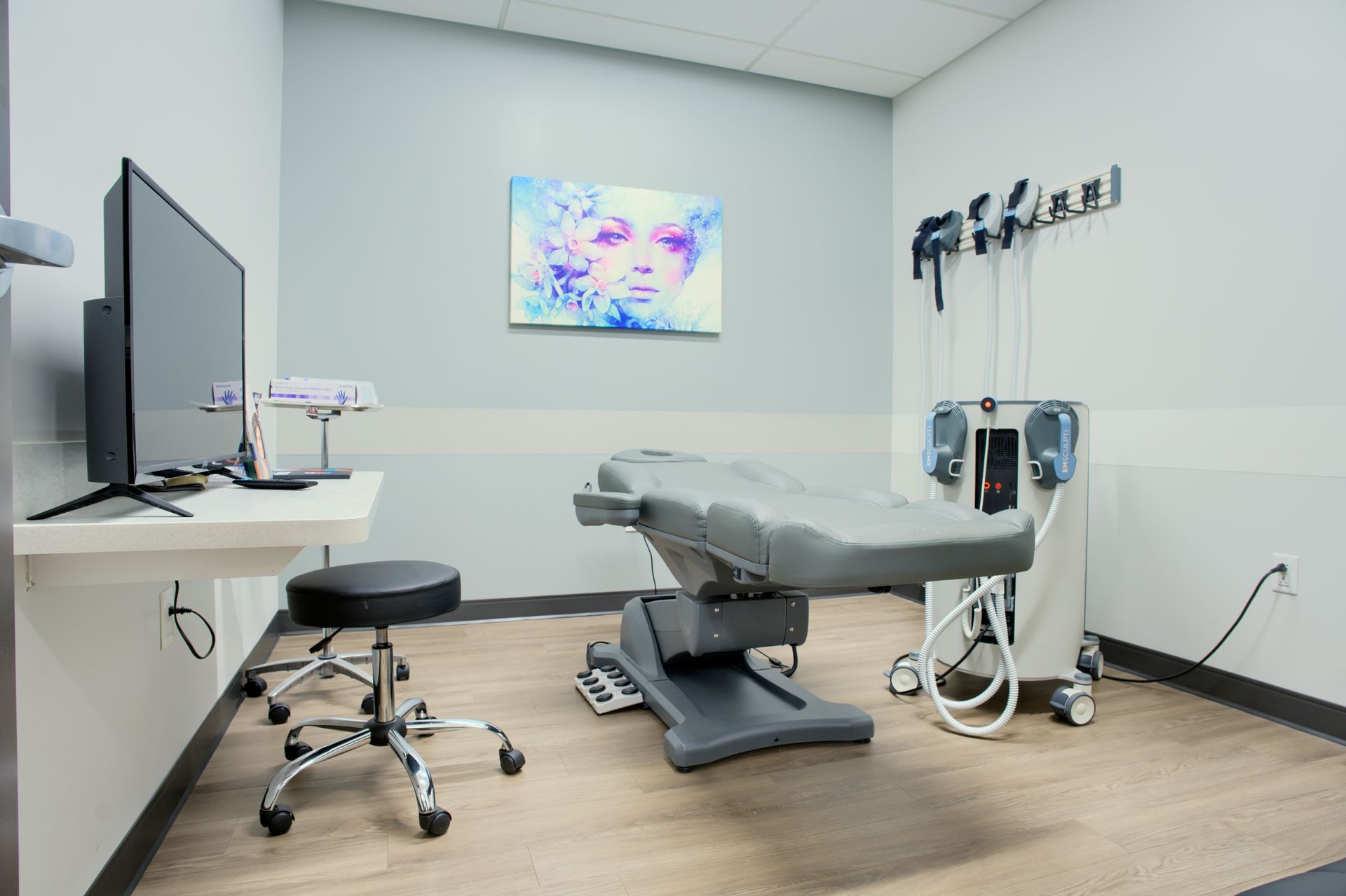 A surgery room with a bed , stool , television , and painting on the wall.
