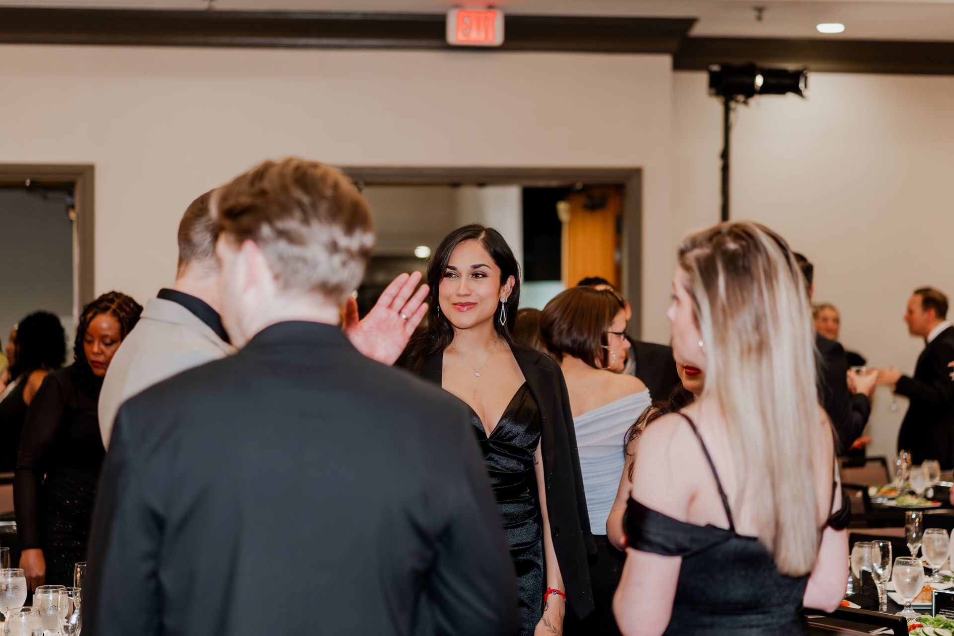 People gather at an indoor event, talking and interacting in a semi-formal setting with tables and guests in the background.