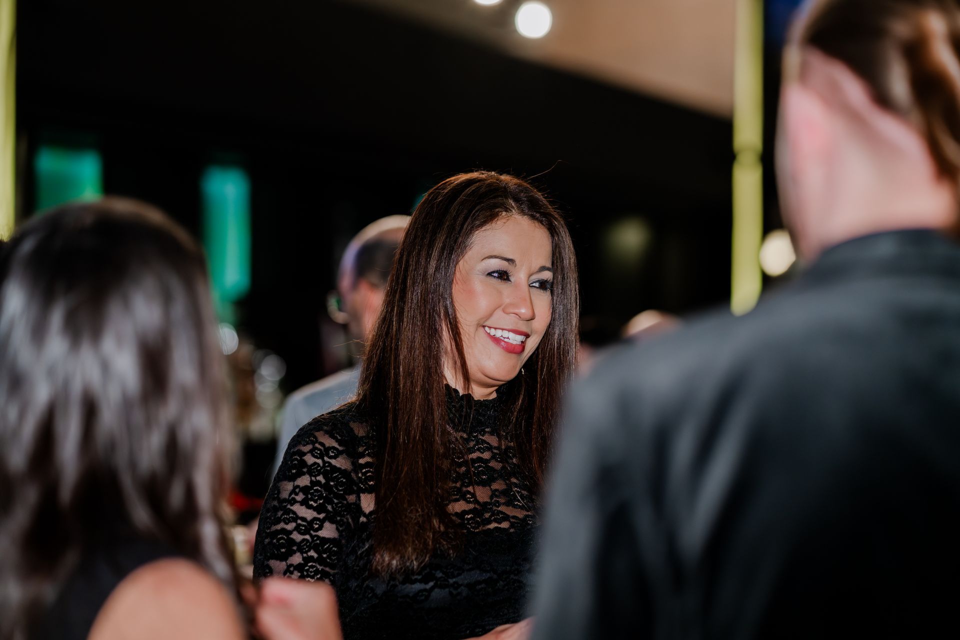 A woman in a black lace top smiling while speaking to people at an indoor event.