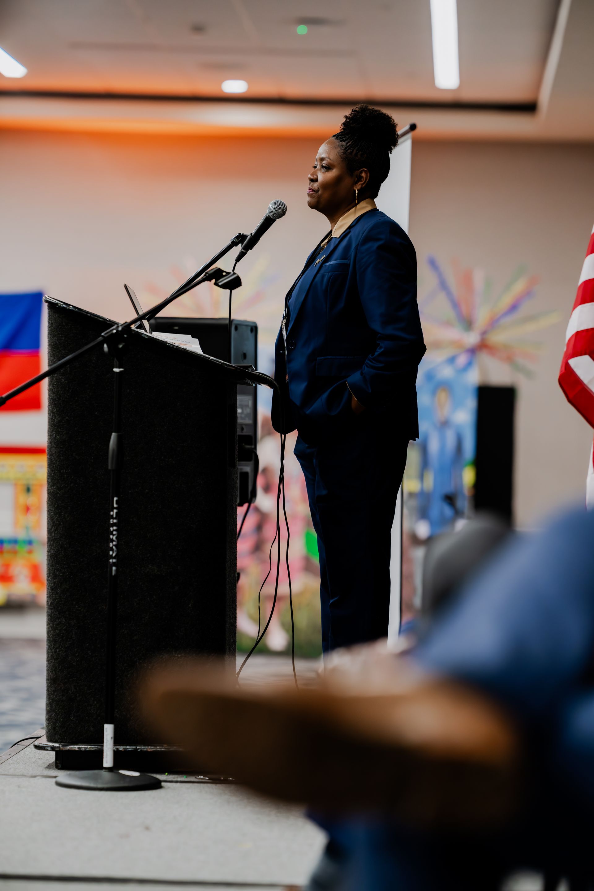 A person in a dark suit stands at a podium with a microphone, addressing an audience in a room with flags.