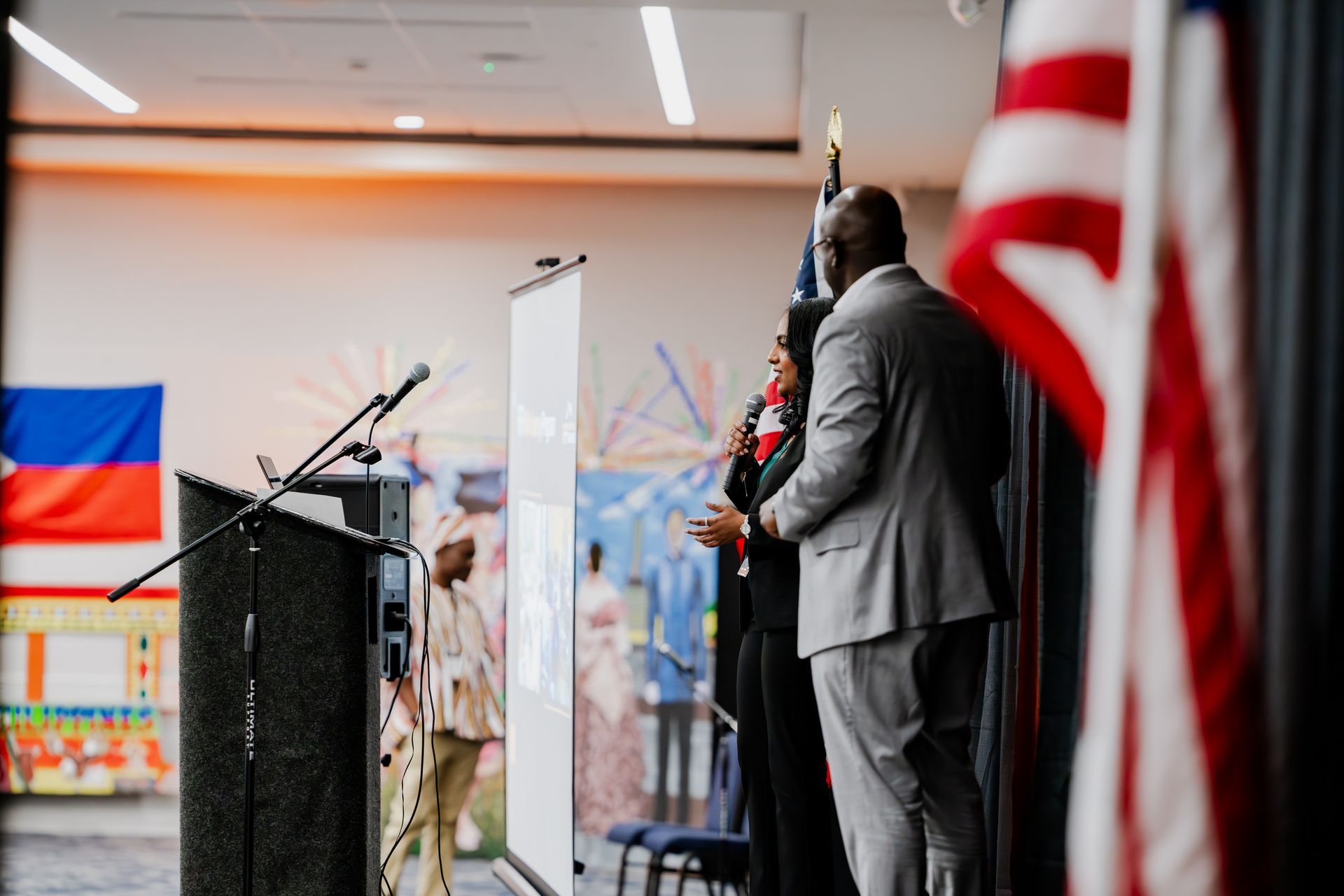 Two people stand at a podium in a hall with flags, including the Haitian flag, on display in the background.