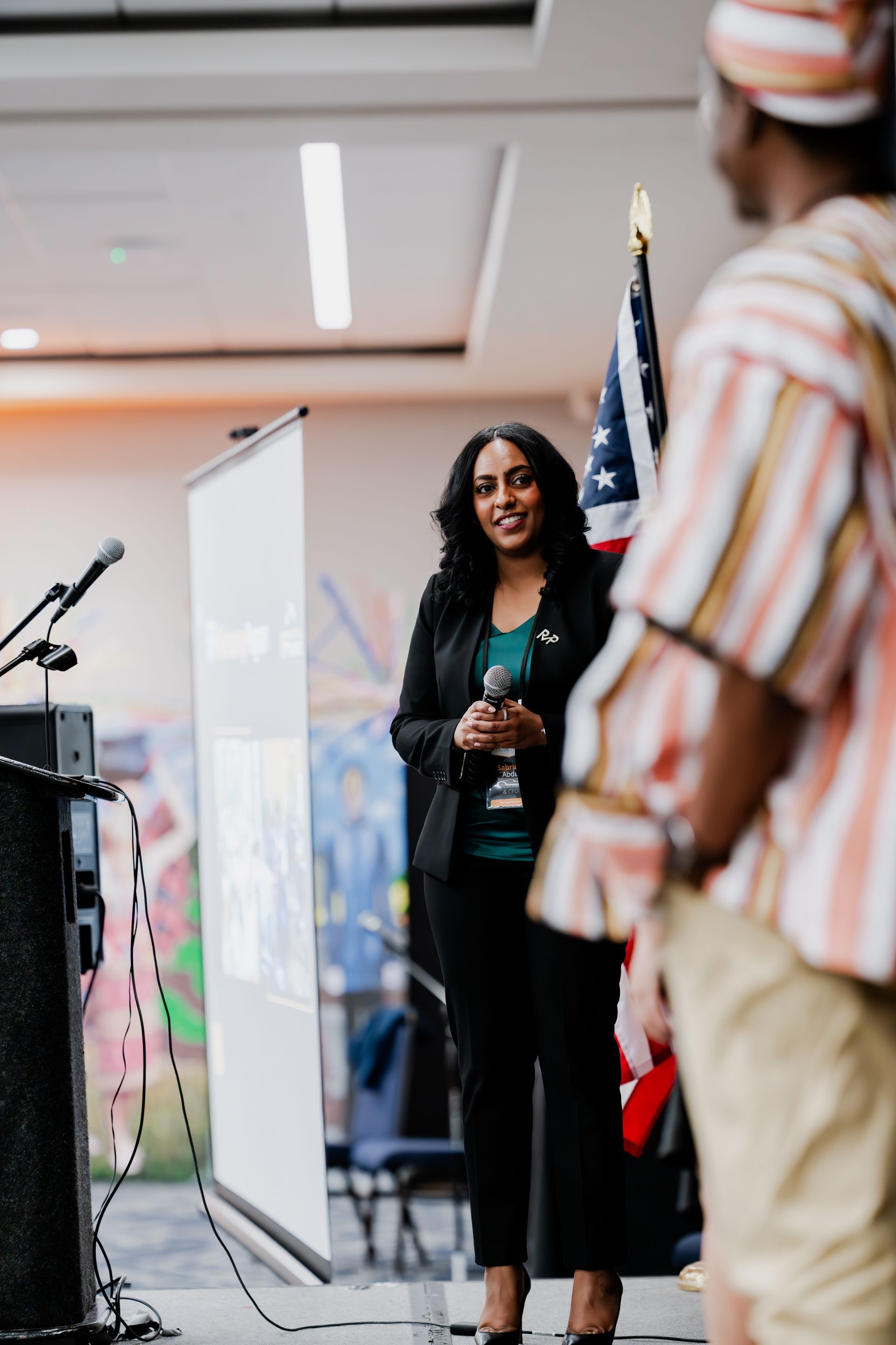 A speaker stands before a projector screen and an American flag, facing an audience member in a striped tunic.