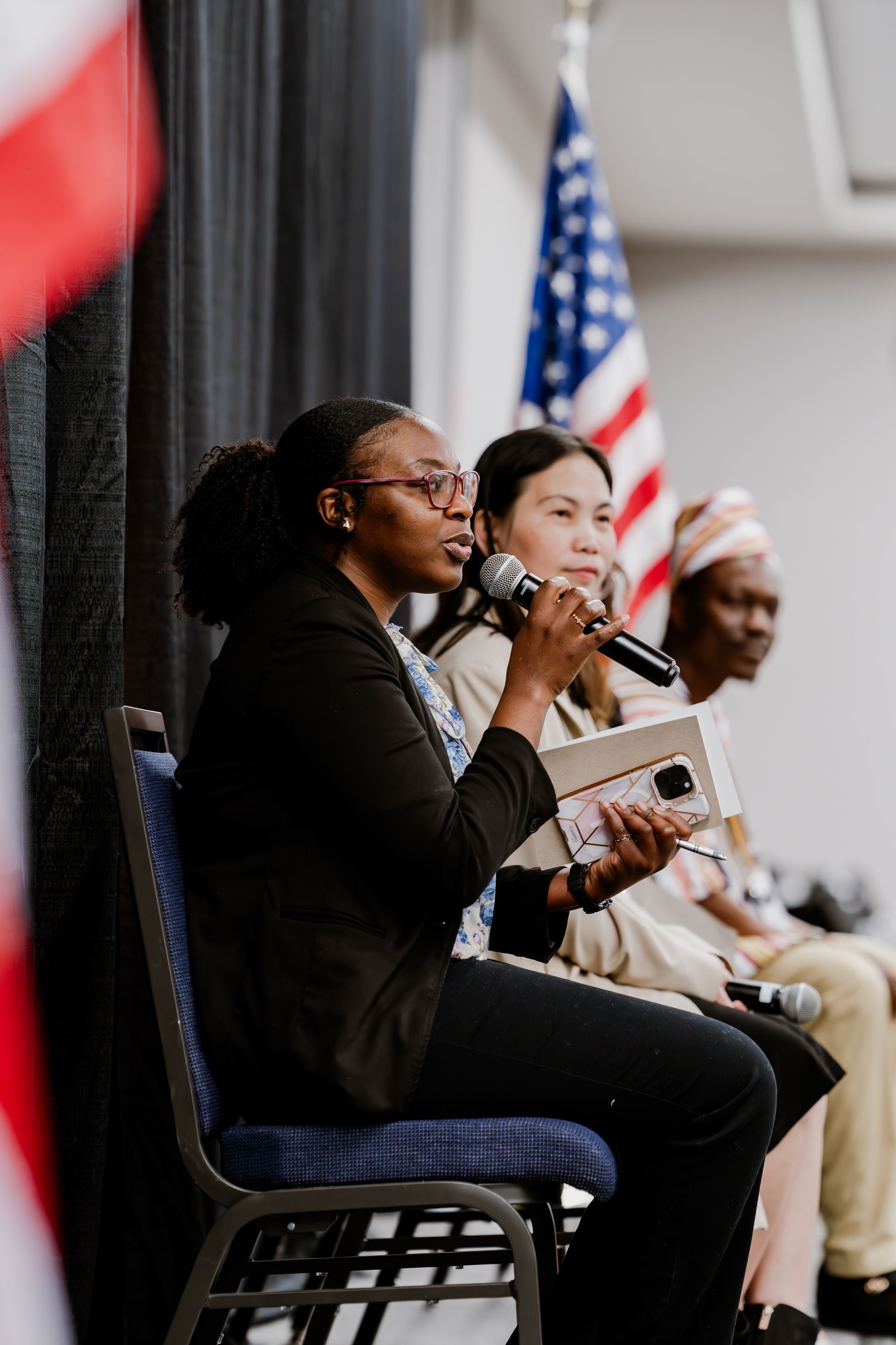 Three people seated on a panel during an event, with the speaker in the foreground holding a microphone and a notepad.