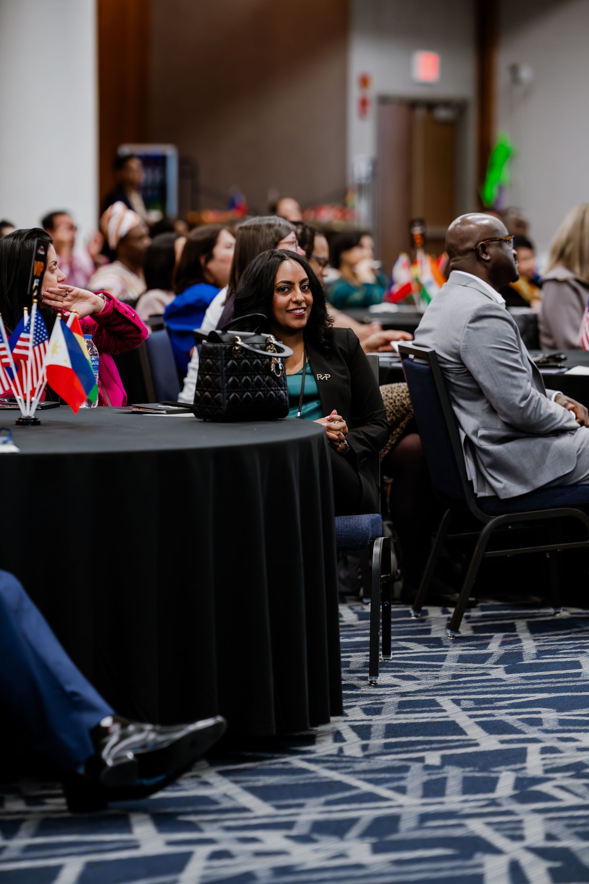 A woman smiles while seated at a round table during a conference, with attendees in a blurred background.