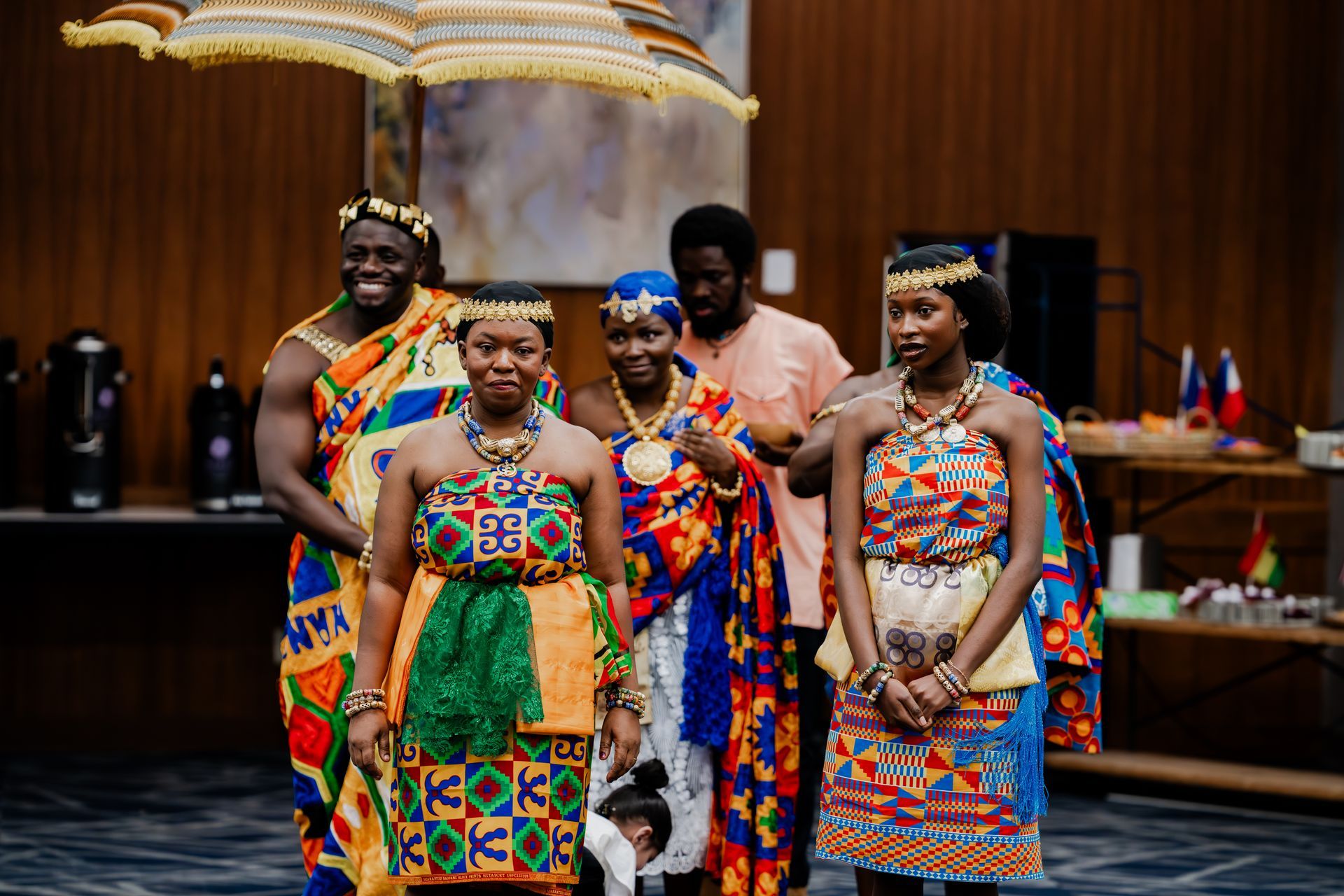 A group of people wearing traditional, colorful Kente cloth garments and jewelry stands together indoors.