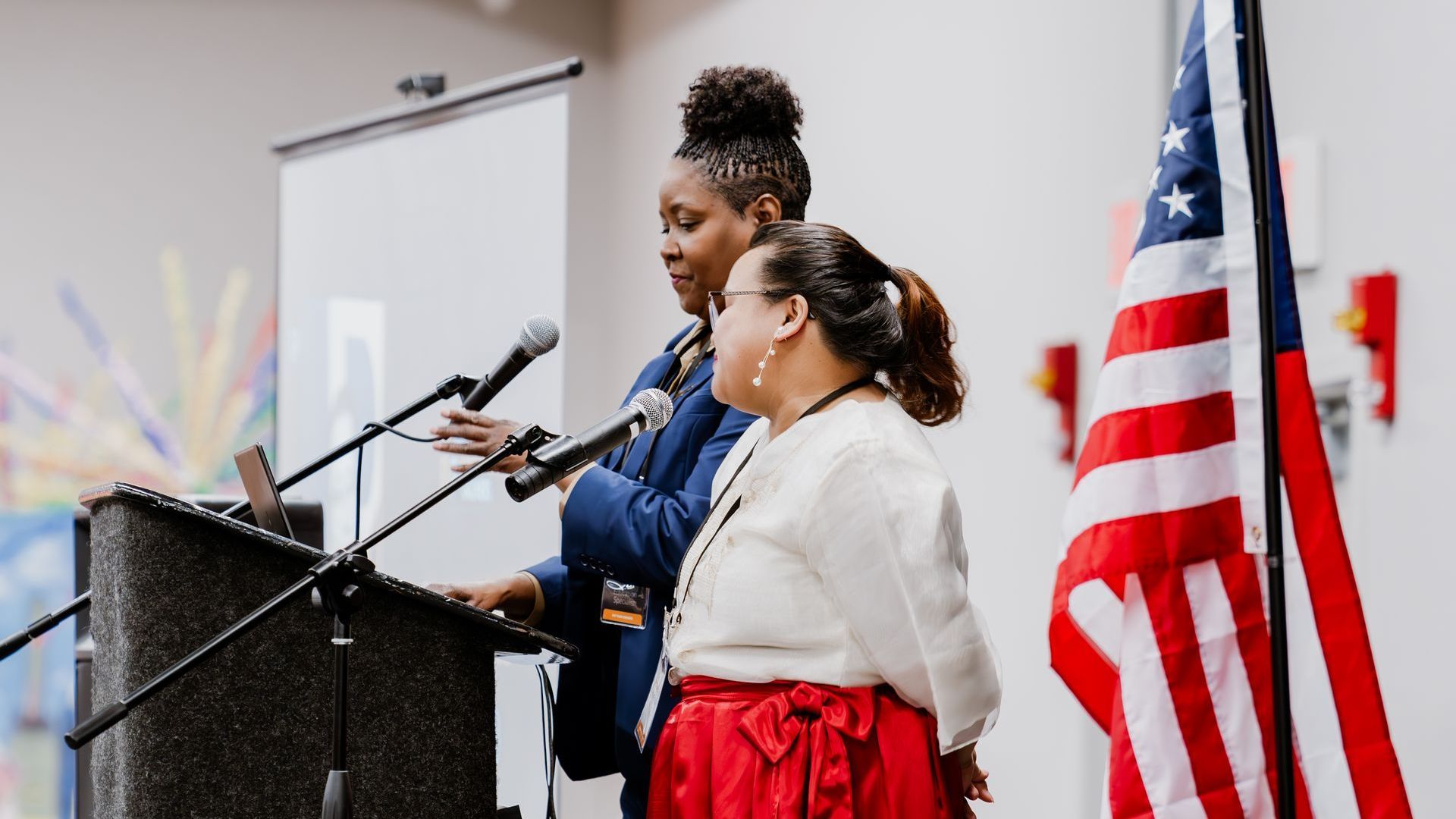 Two women speaking at a podium; American flag in background.