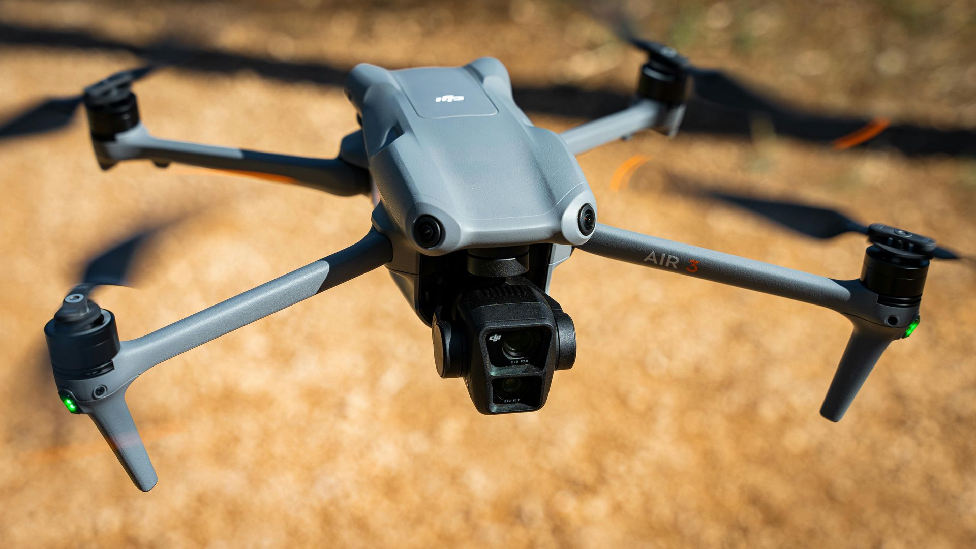 Grey DJI Mavic 3 drone with propellers extended, hovering over brown ground.