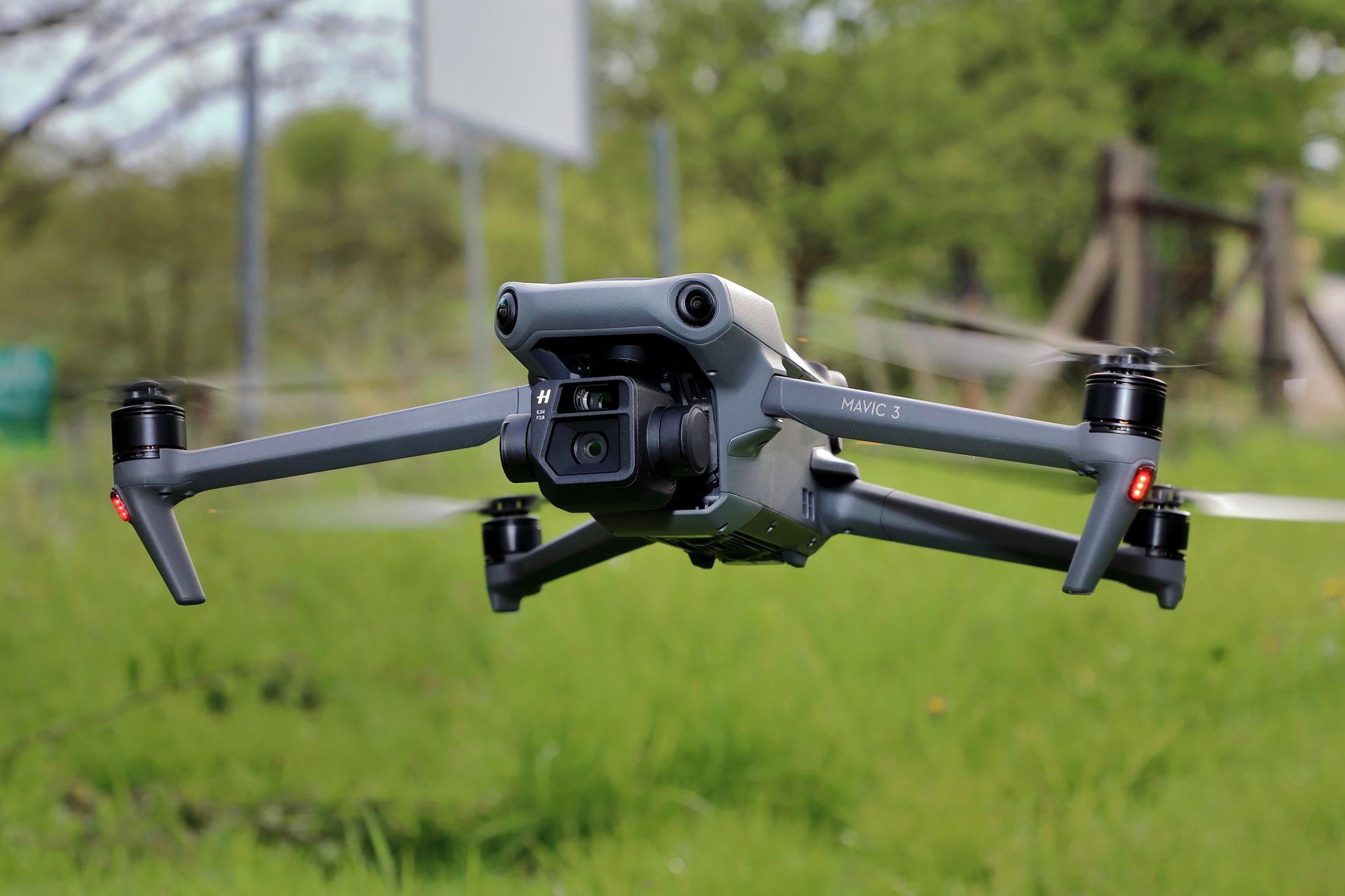 Grey drone in flight over green grass with a blurry background of trees and sign.