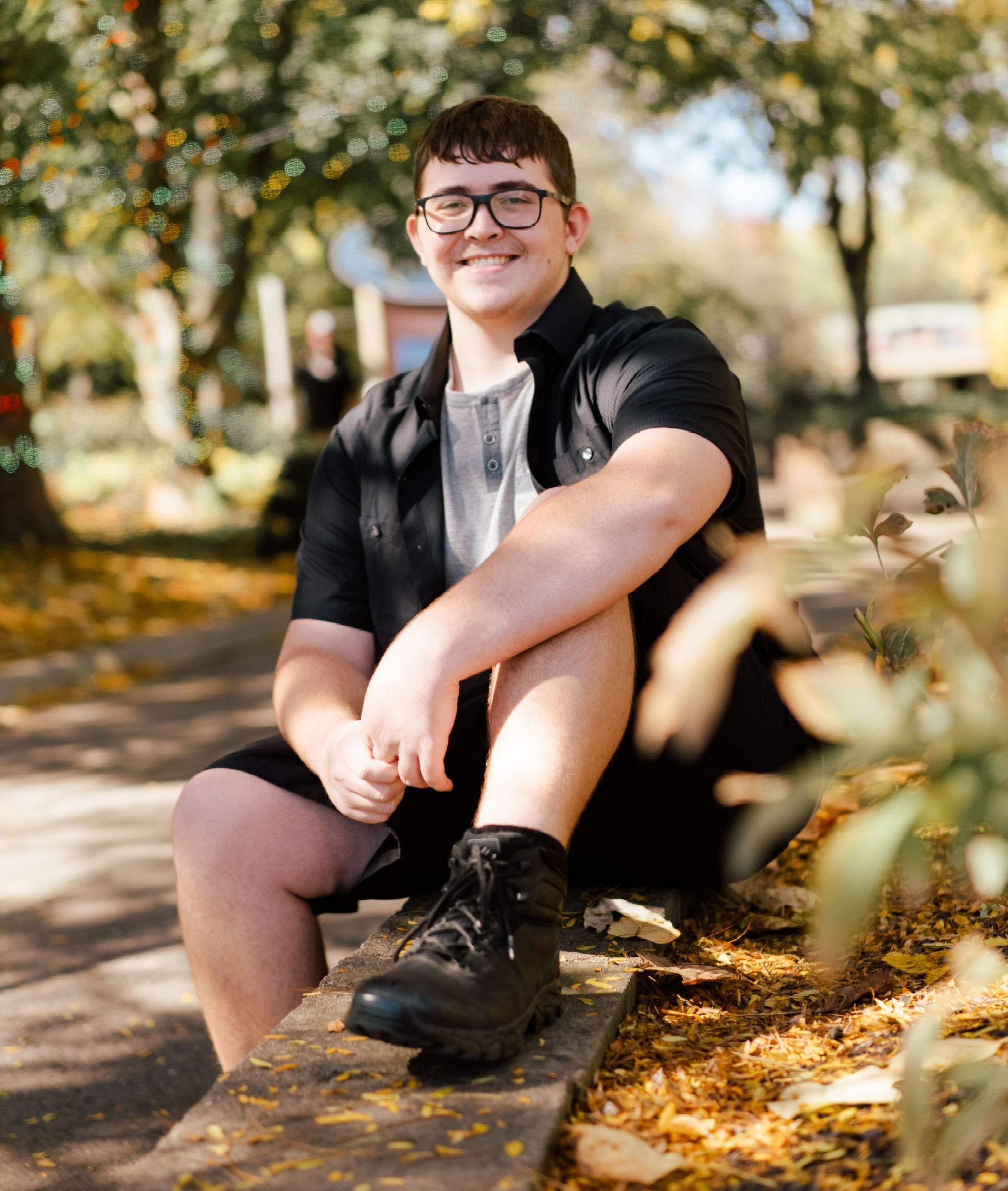 High school senior wearing glasses is sitting on a bench in a park .