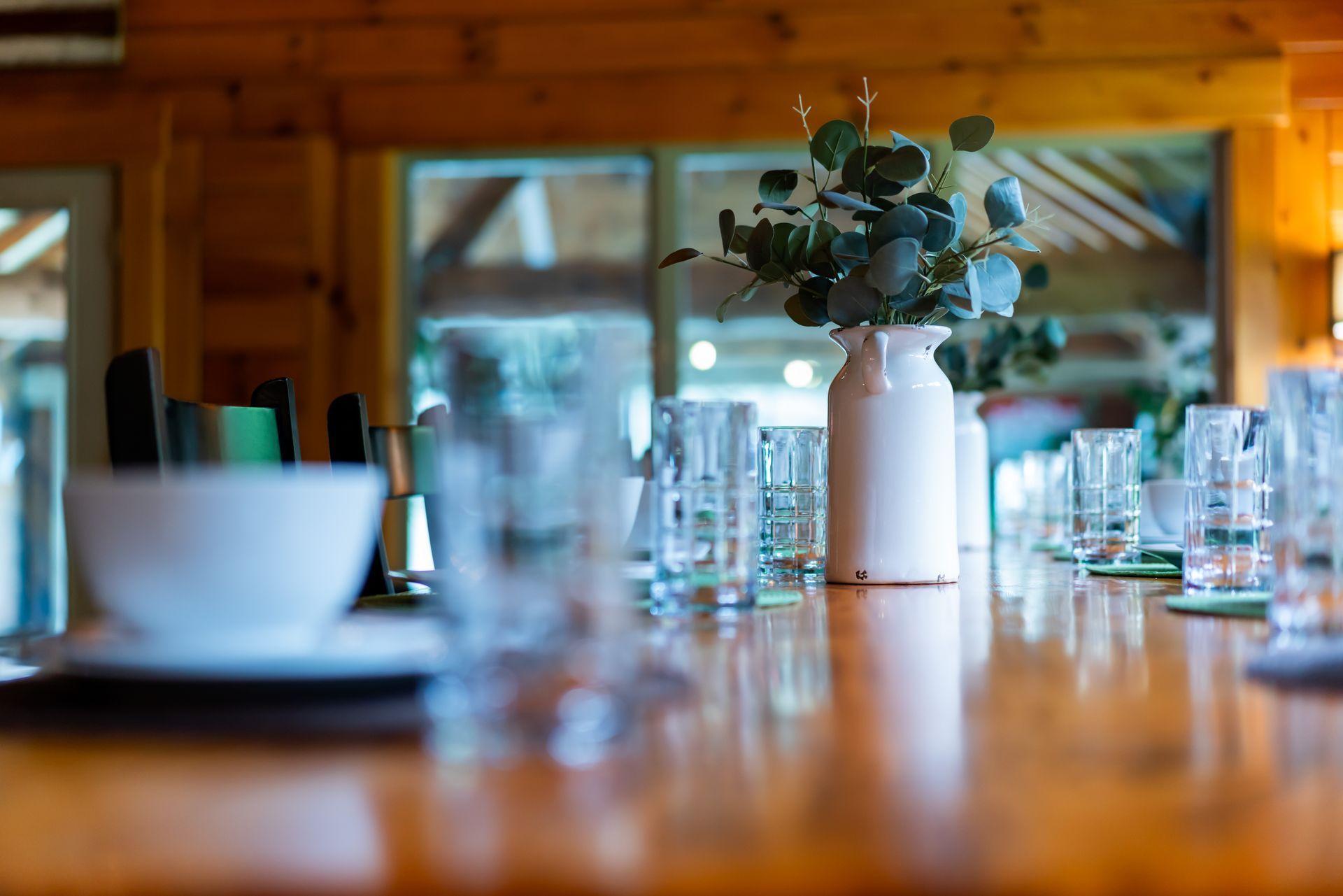 a long wooden table with plates , bowls , glasses and a vase of flowers on it .