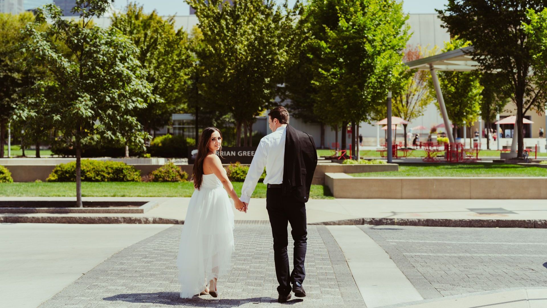 Bride and groom walking hand-in-hand in a sunny park; the bride wears a white dress and the groom a black suit.