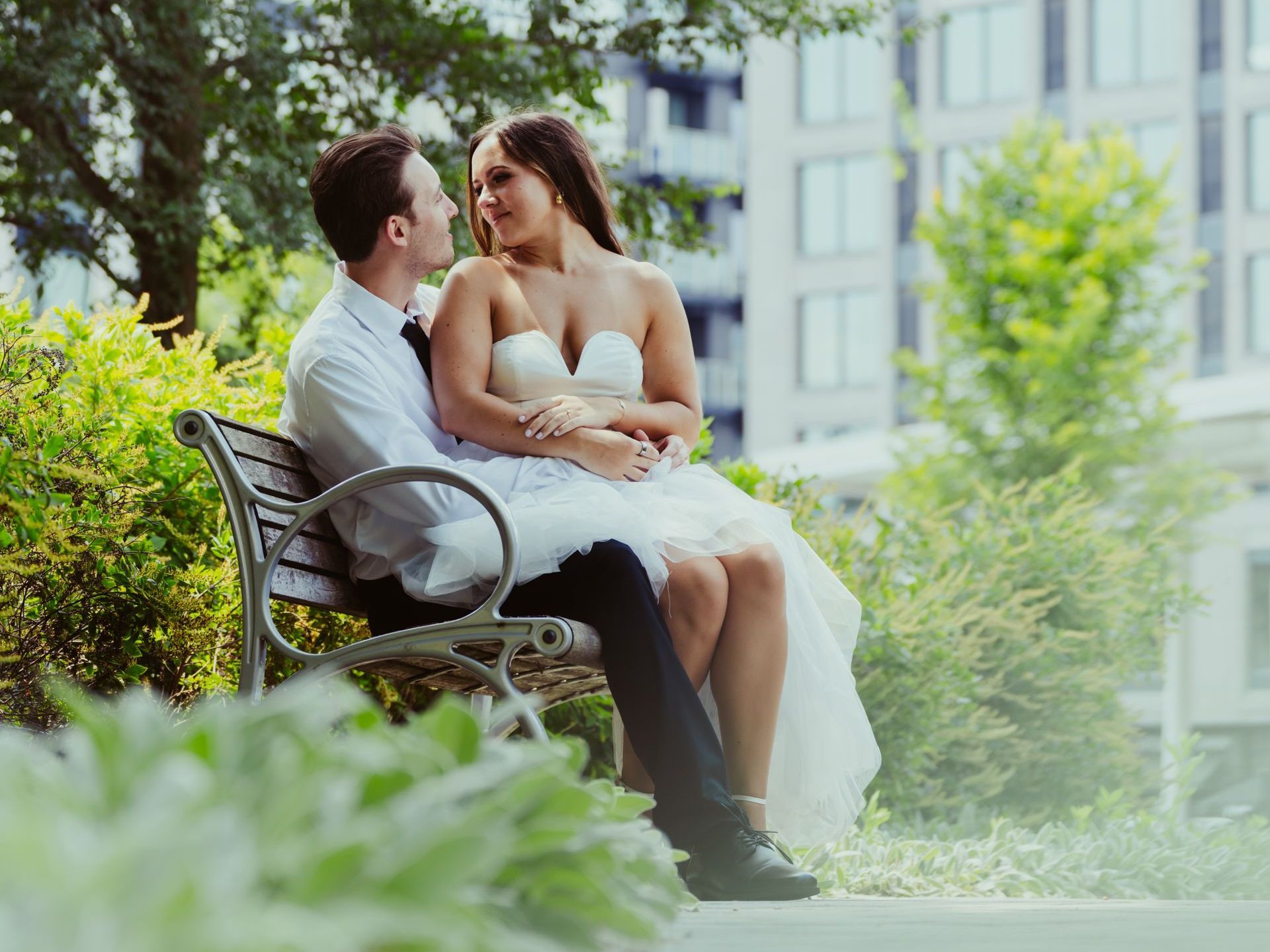 Couple in wedding attire embrace on a park bench, looking at each other.