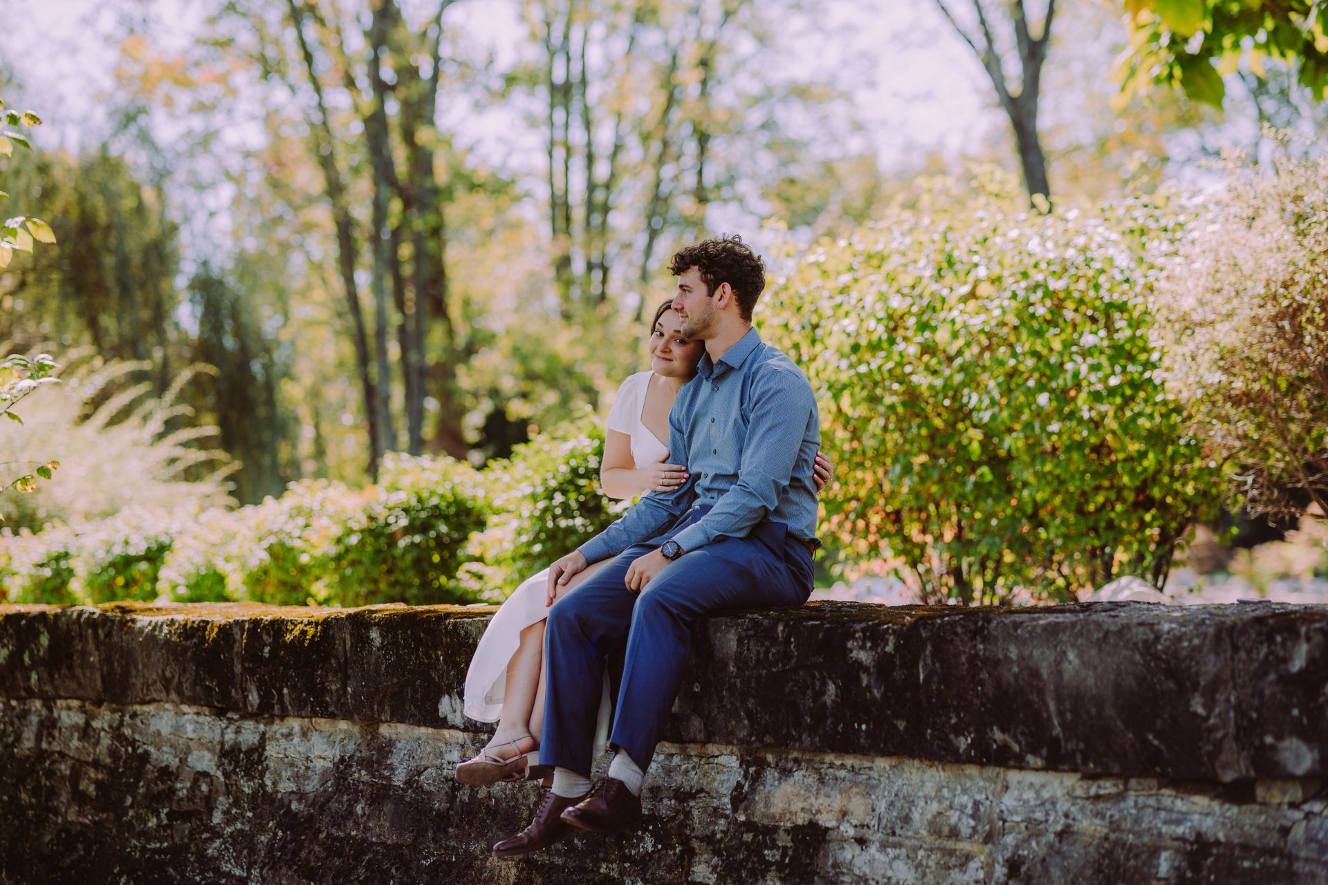 Couple sits close on a stone wall, embracing. Bright, sunny outdoors setting with green trees and bushes.