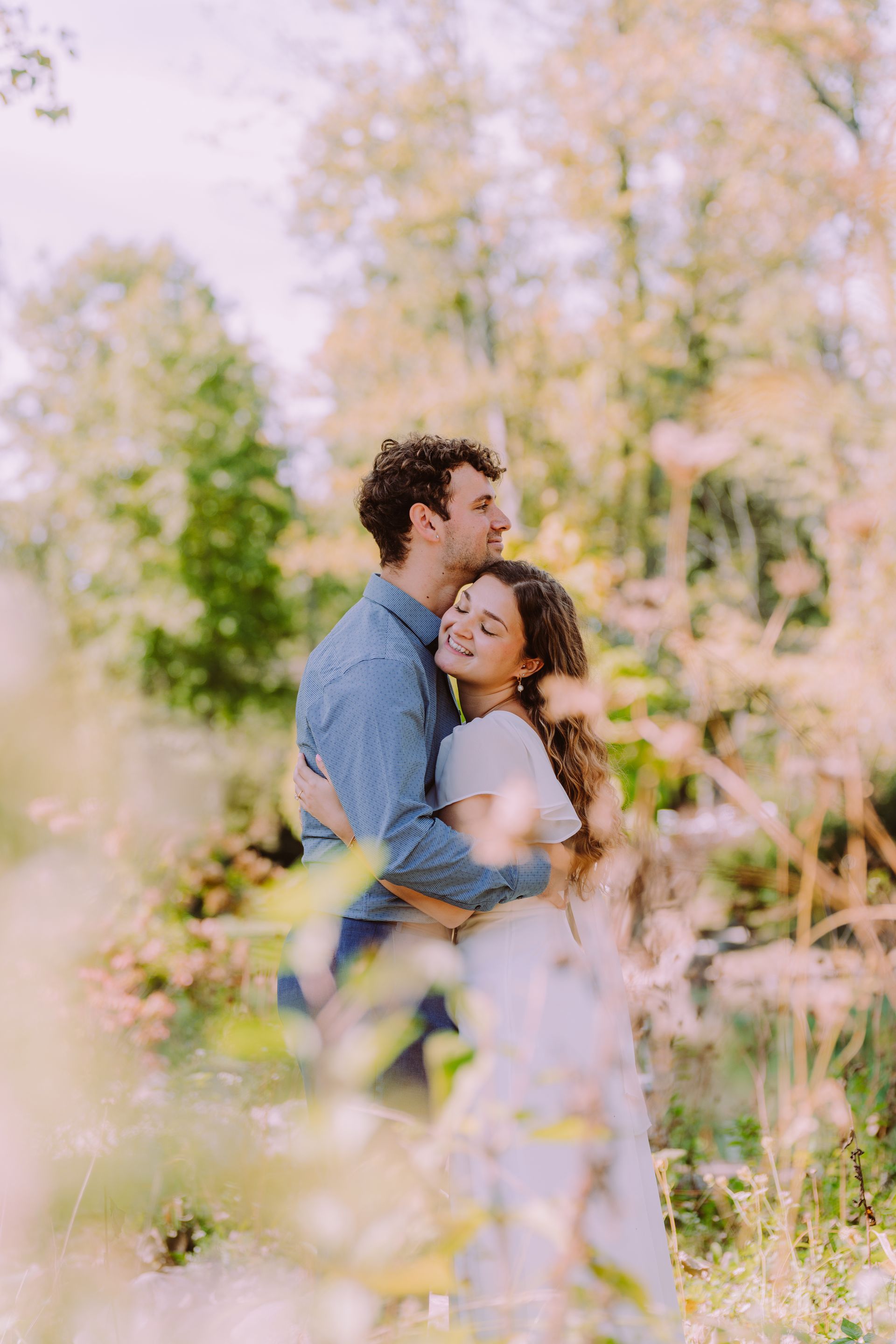 Couple embracing in a field of wildflowers; man hugs woman from behind, both smiling near trees.