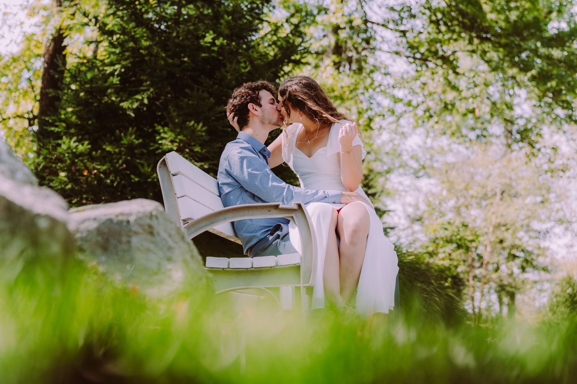 Couple kissing on a park bench, sunlight, trees, and greenery in the background.