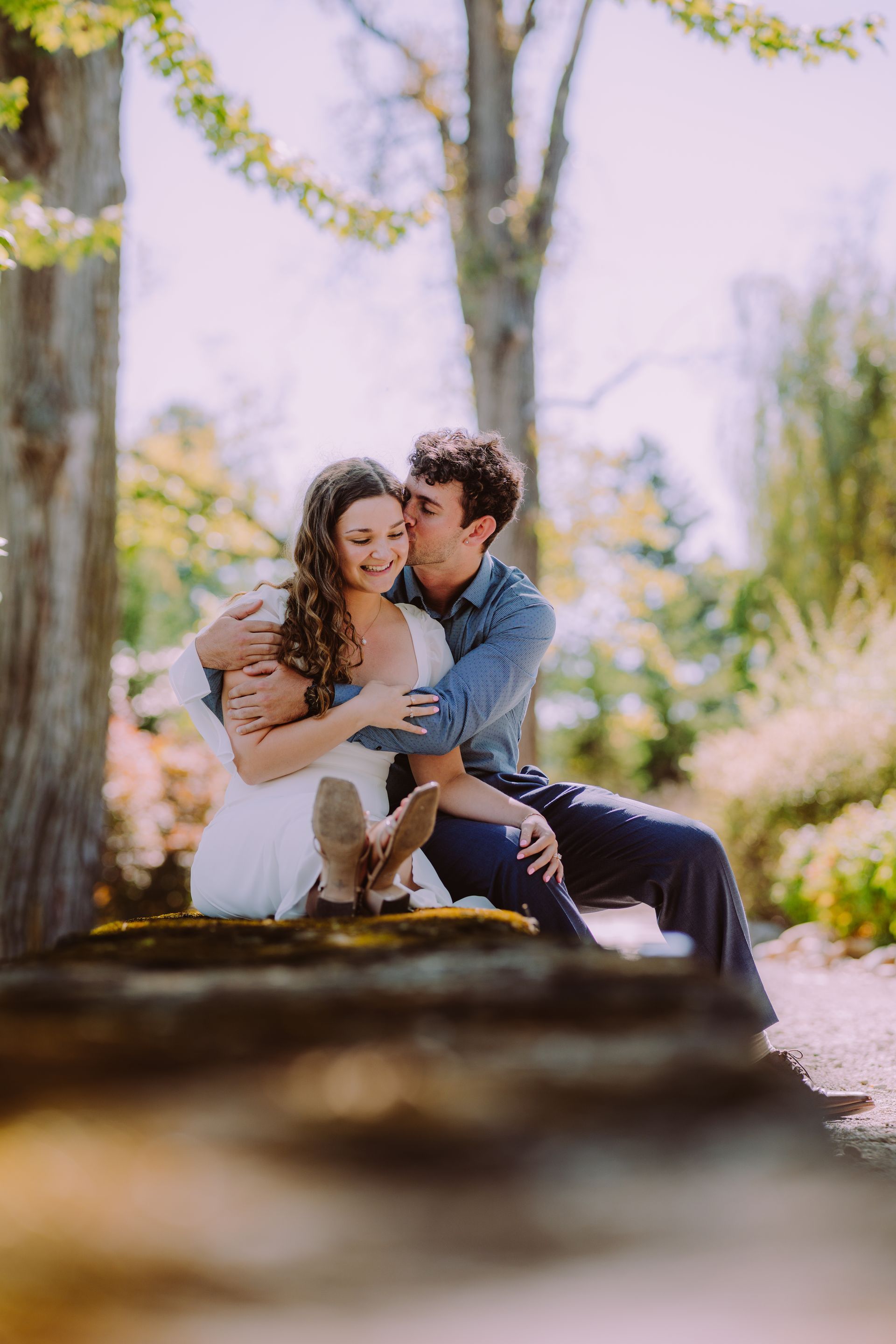 Couple hugging and kissing, sitting outdoors under a tree, with sunny background.